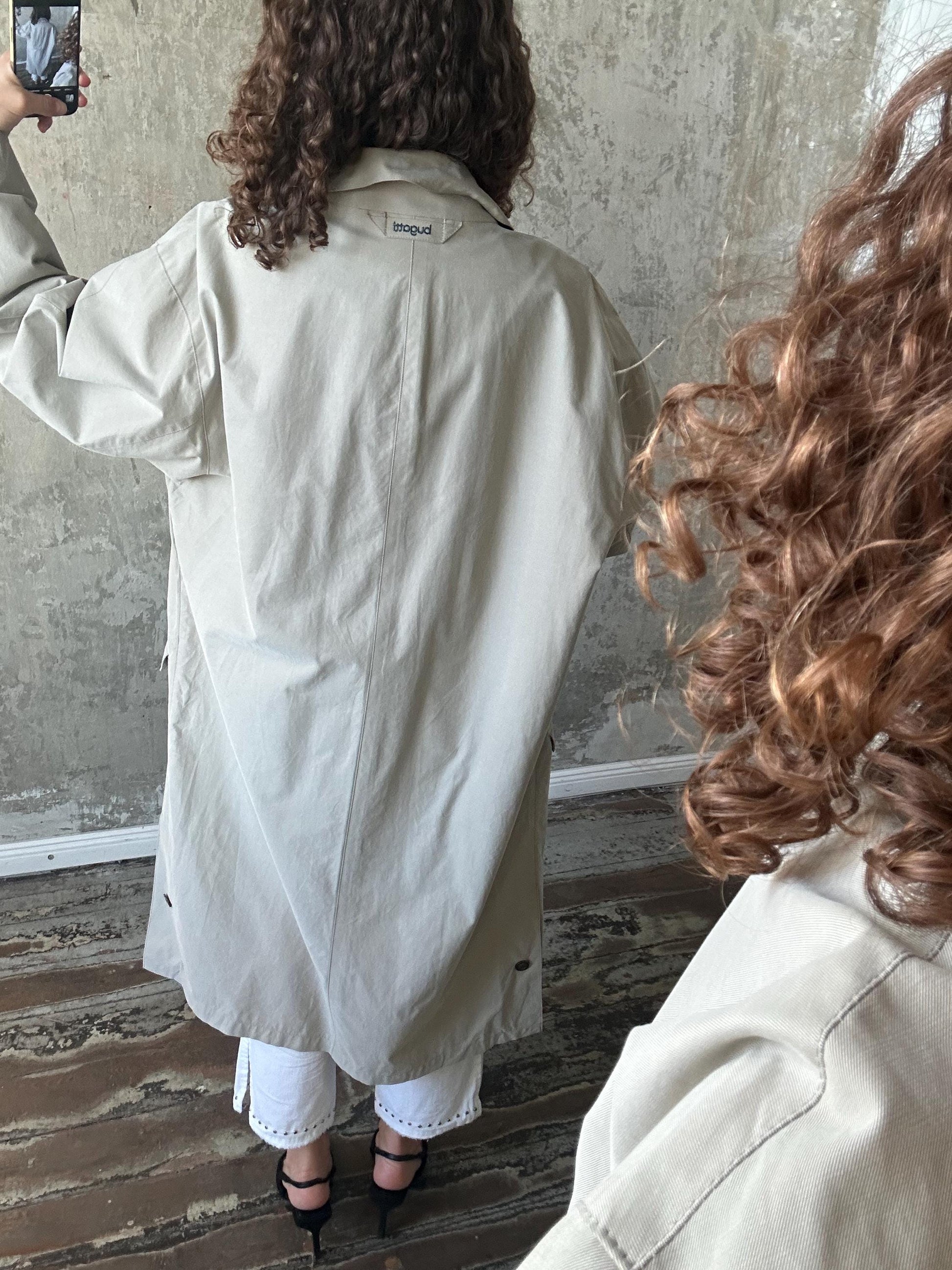 a person wearing a beige trench coat, taking a selfie with a cell phone.