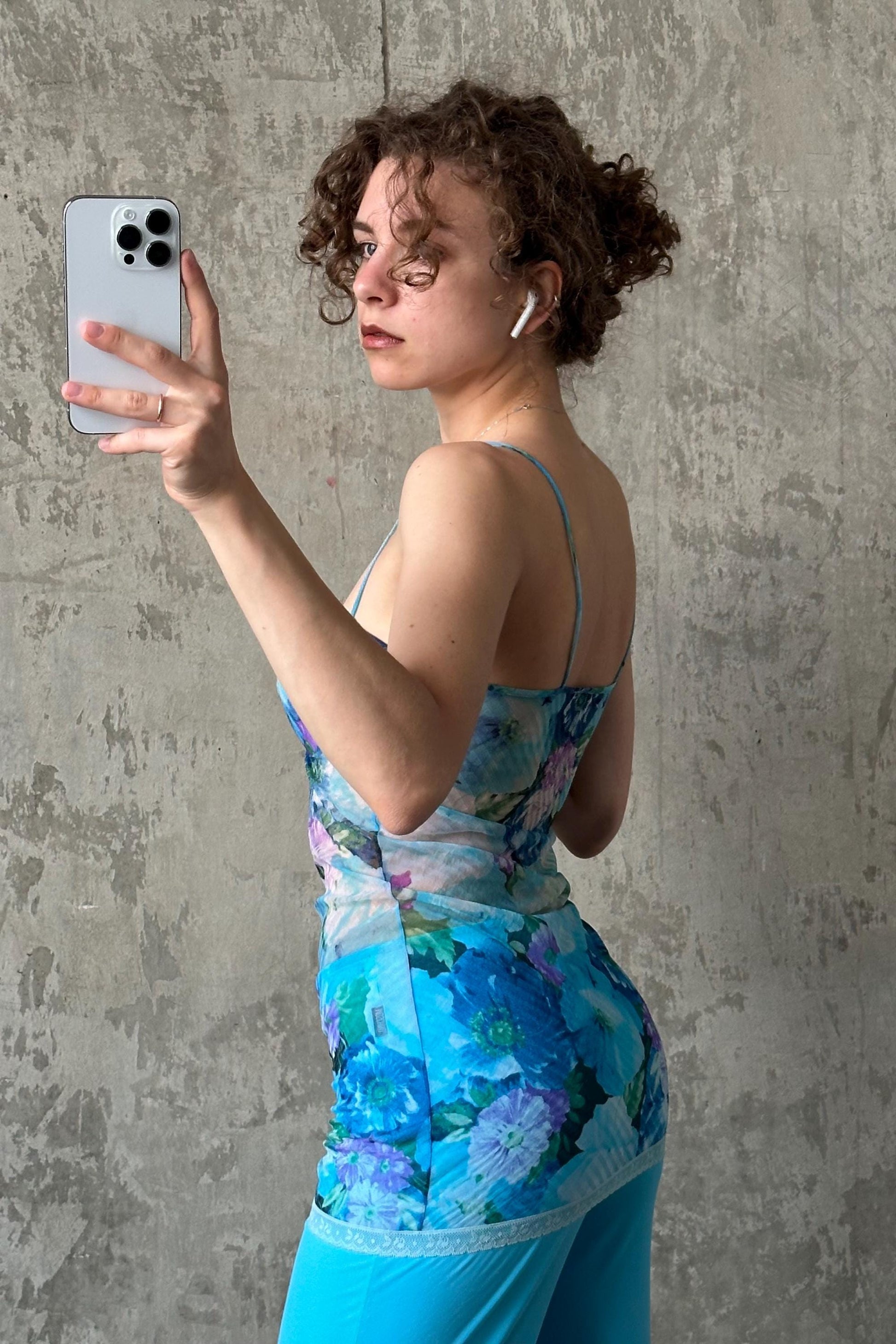 A woman with curly hair is taking a selfie with a smartphone, wearing a blue floral dress.