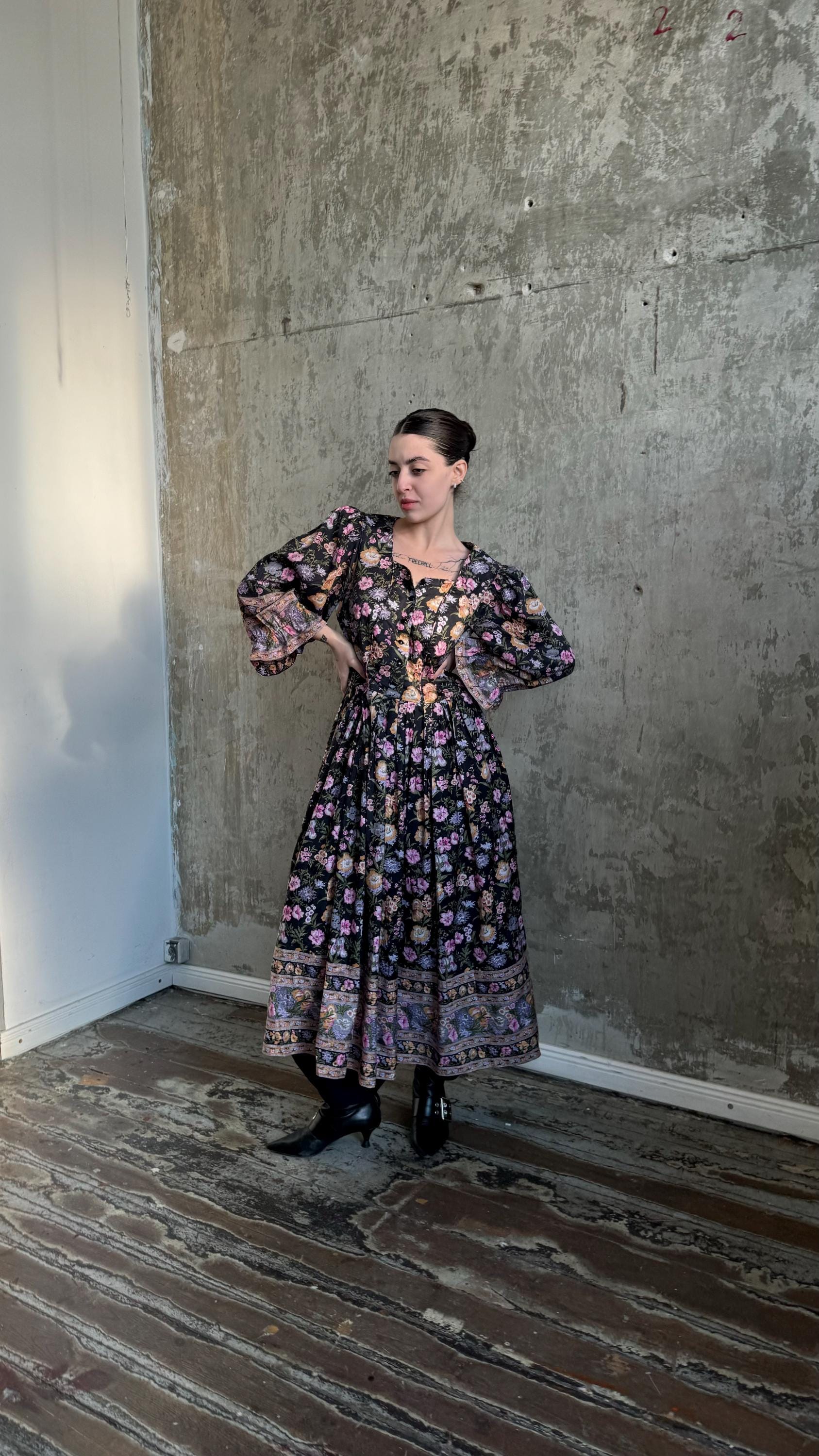 A woman wearing a long, floral-patterned dress stands in a room with a concrete wall.