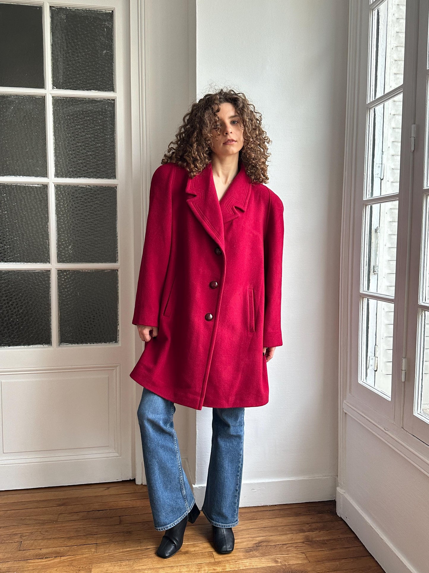 A person wearing a red coat and blue jeans stands in a room with a window.