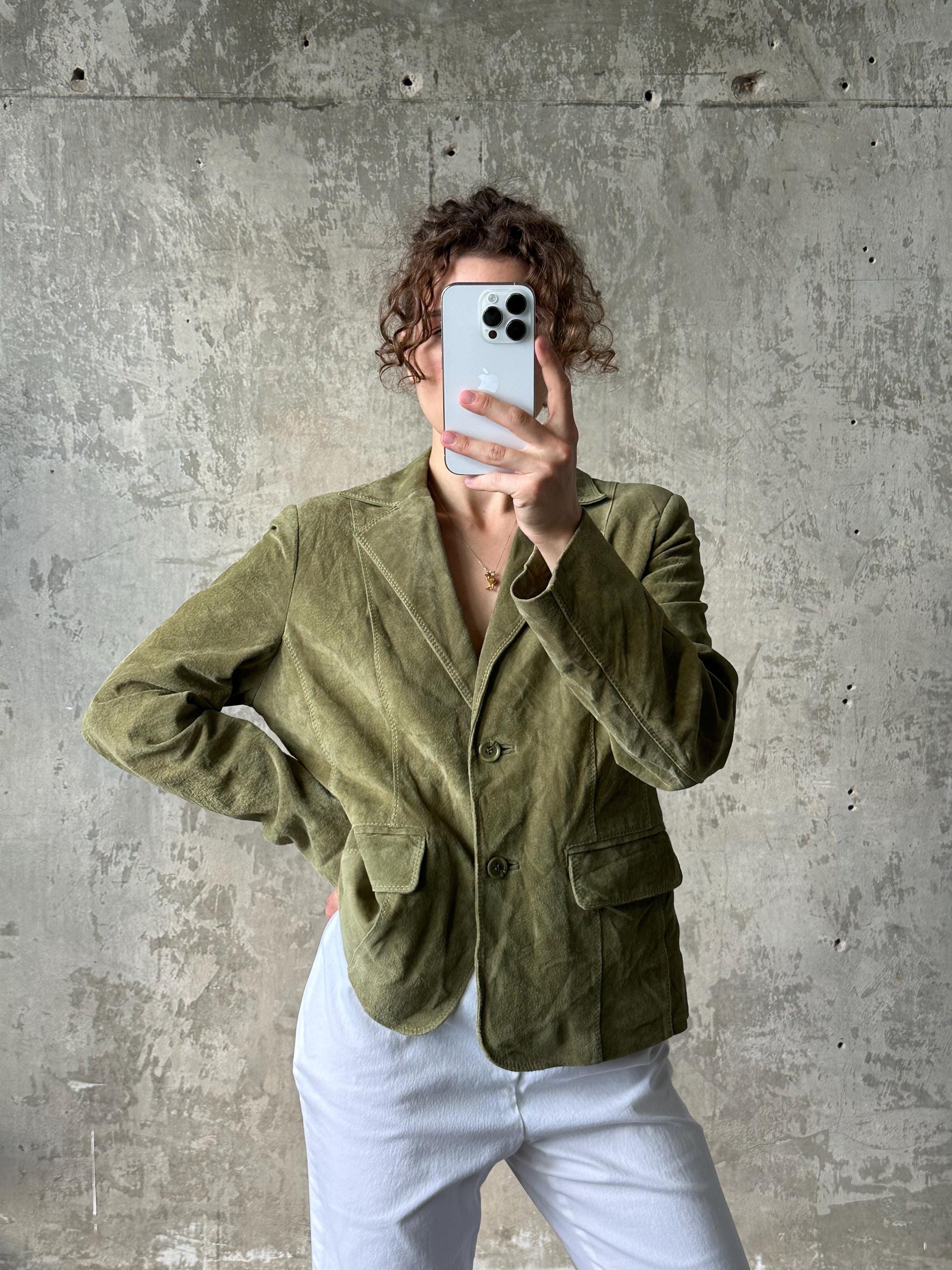 A person is taking a selfie with a white smartphone, wearing a green blazer and white pants against a concrete wall background.