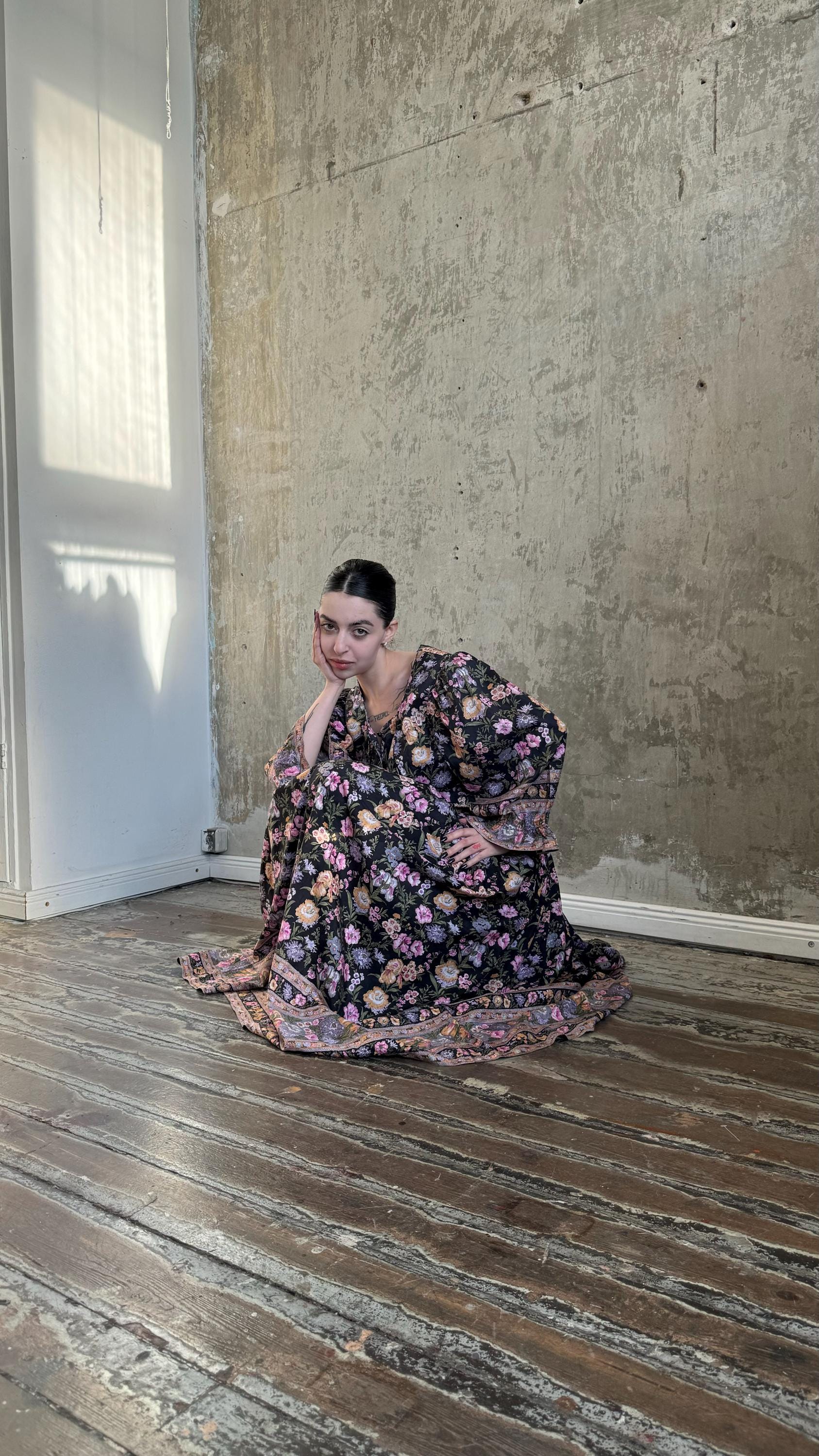 A woman in a floral dress sits on a wooden floor, her hand resting on her chin in a thoughtful pose.