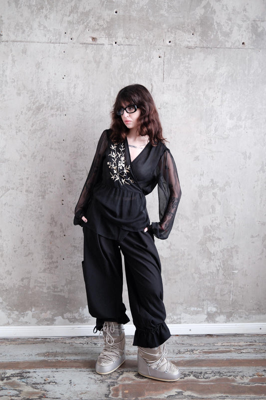 A woman in a black outfit with sheer sleeves stands against a plain white wall, her gaze directed off to the side.