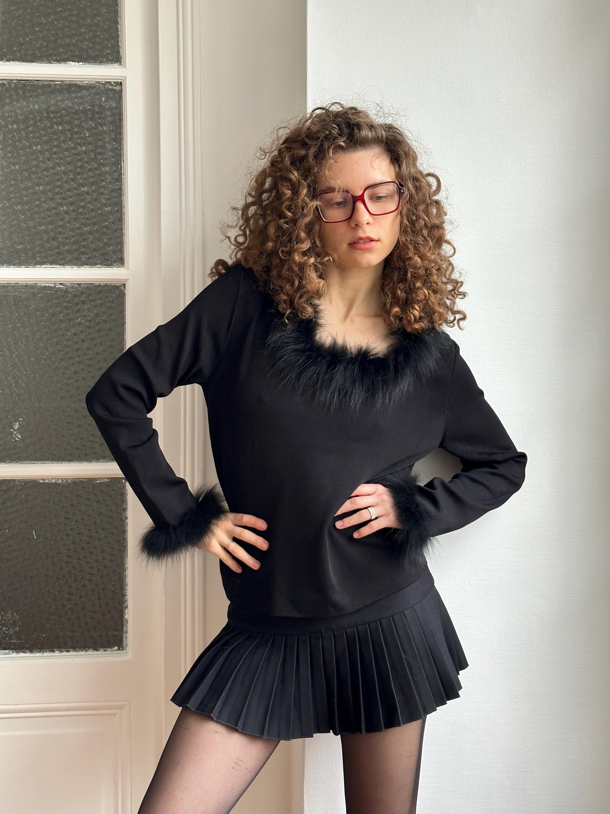 A woman with curly hair and glasses is posing in a black outfit with a fur collar, standing in front of a window.