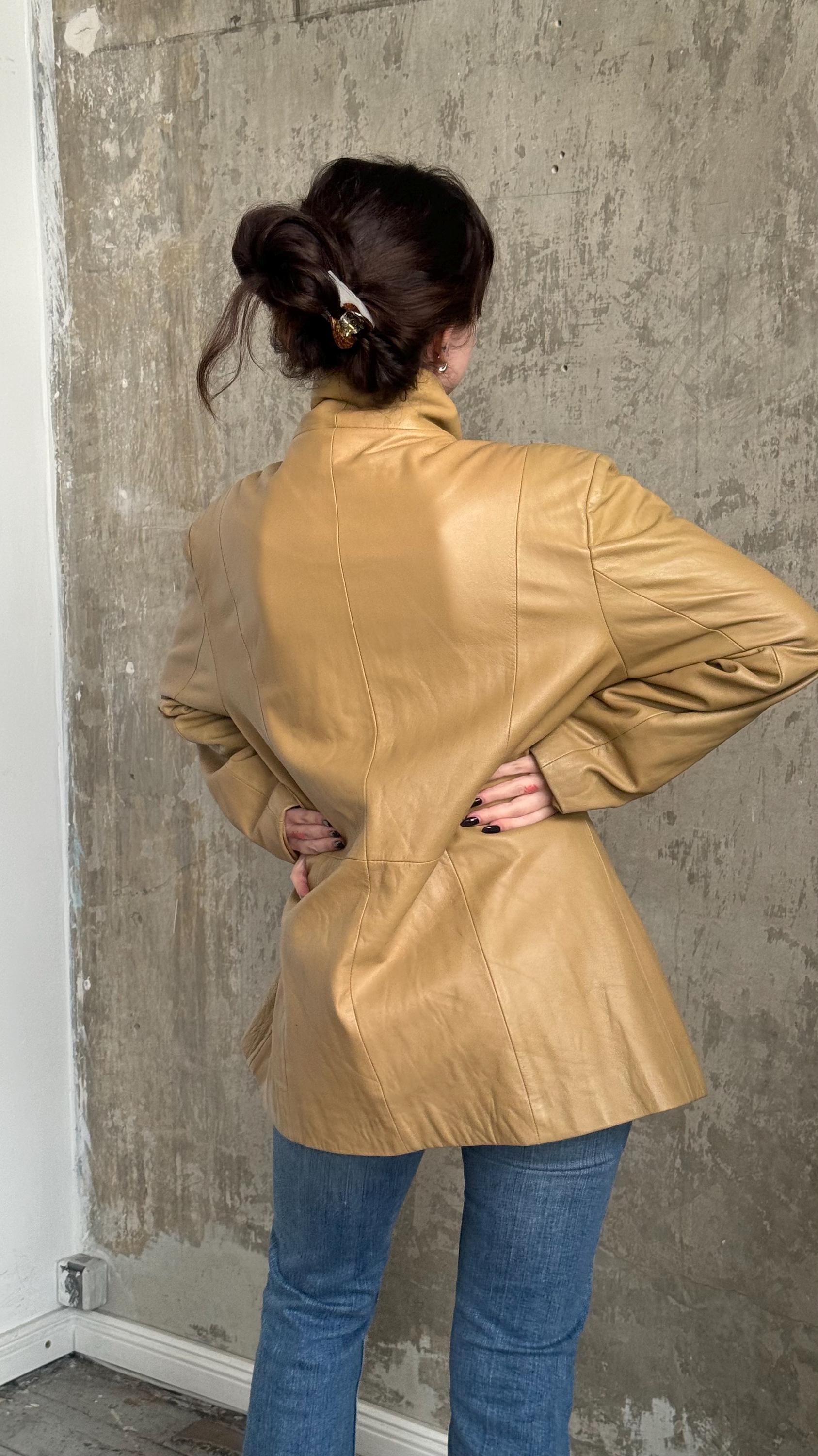A woman wearing a tan leather jacket stands in front of a concrete wall, her back to the camera.