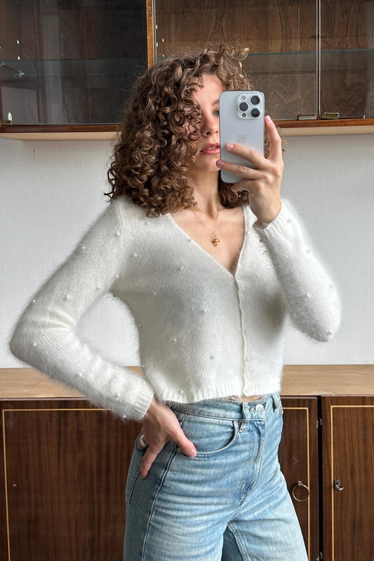 A woman with curly hair is taking a selfie with her iPhone, wearing a white sweater and blue jeans.