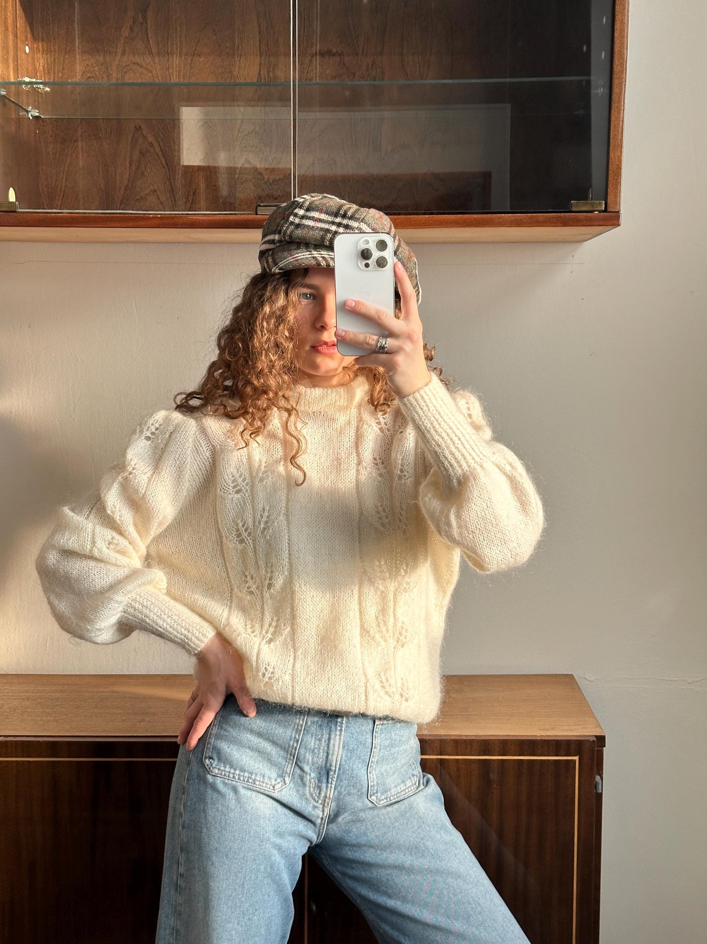 A woman with curly hair is taking a selfie with a white iPhone, wearing a white sweater and blue jeans.