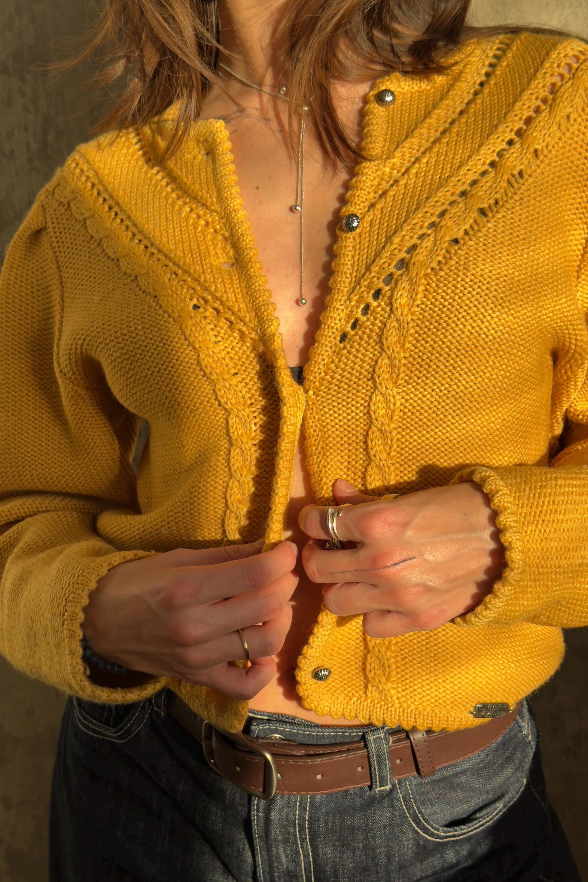 A person wearing a yellow knitted cardigan and jeans is shown adjusting the buttons of the cardigan.