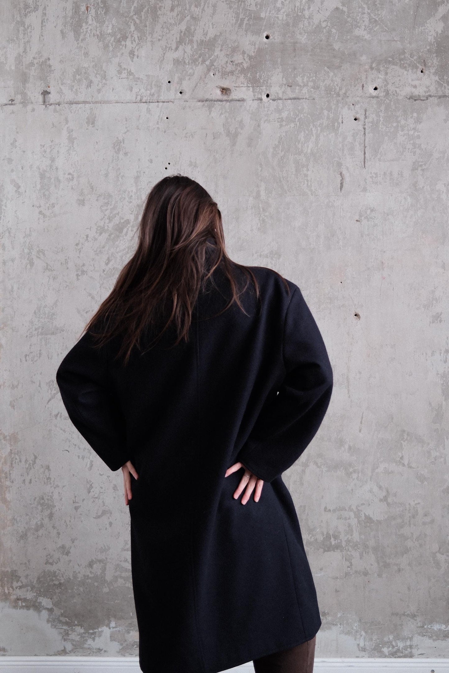 A person with long hair wearing a black coat stands in front of a concrete wall.