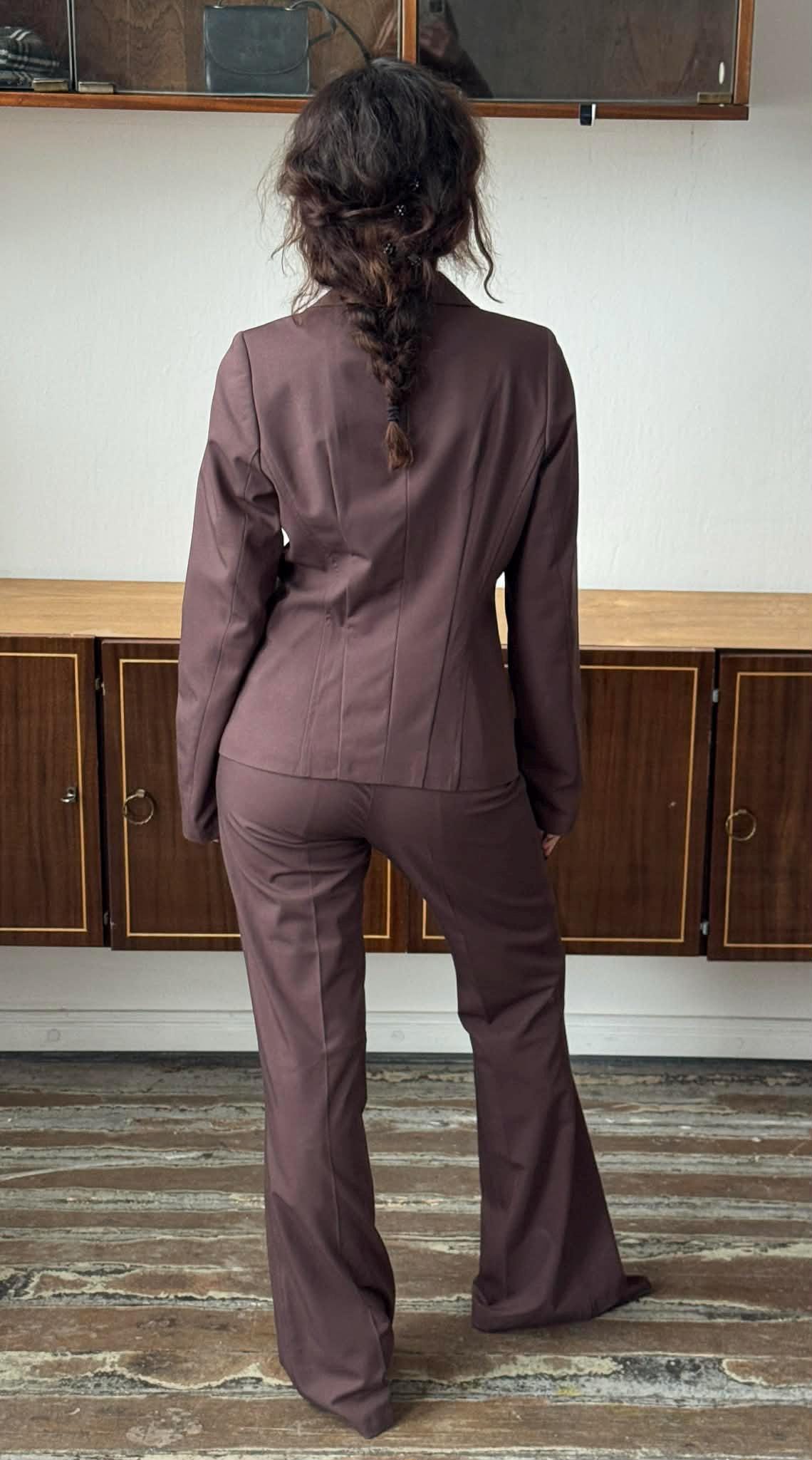 A woman wearing a brown suit stands in a room with wooden cabinets in the background.