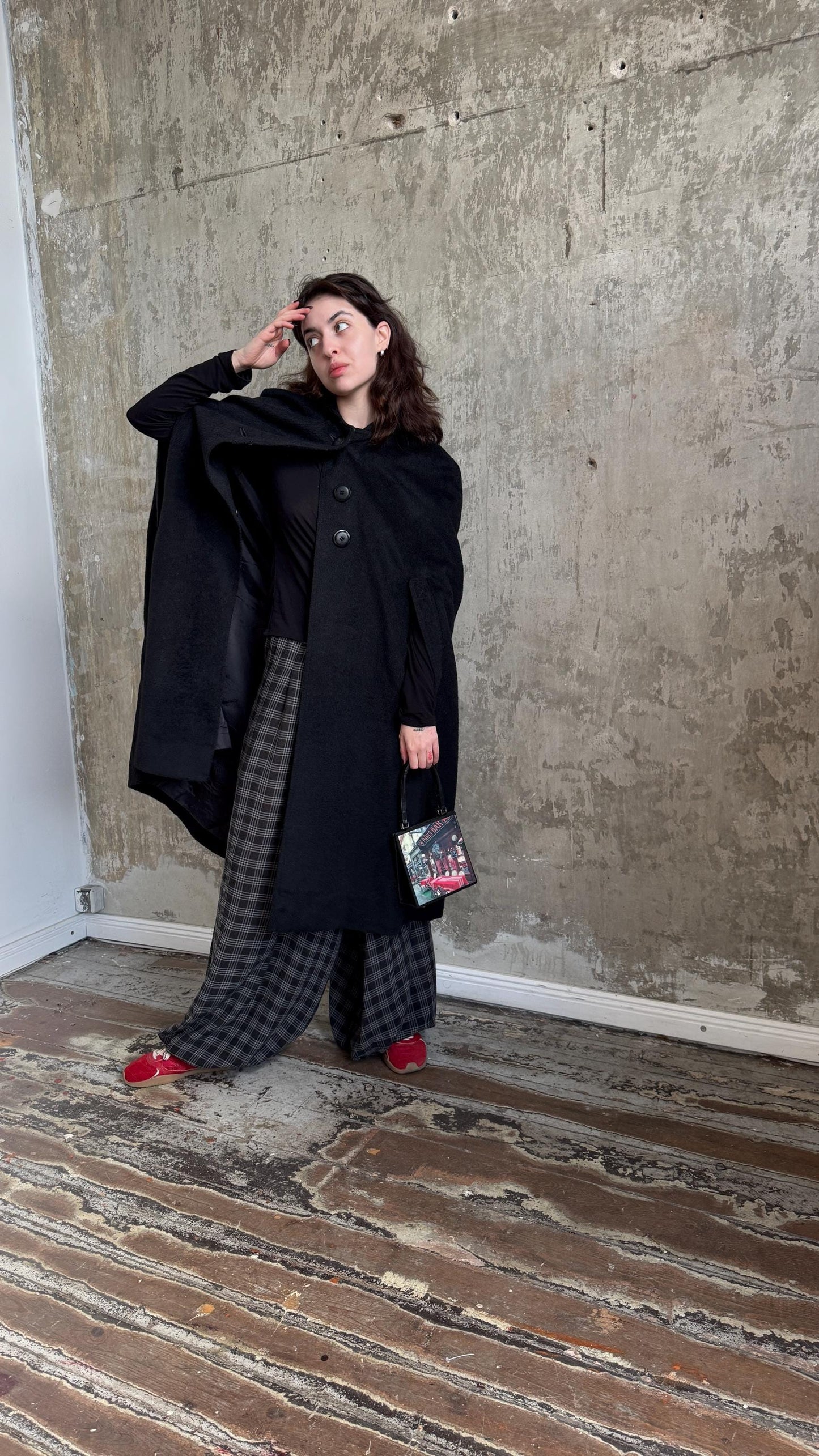 A woman in a black coat and plaid skirt stands in a room with a concrete wall, holding a small bag.
