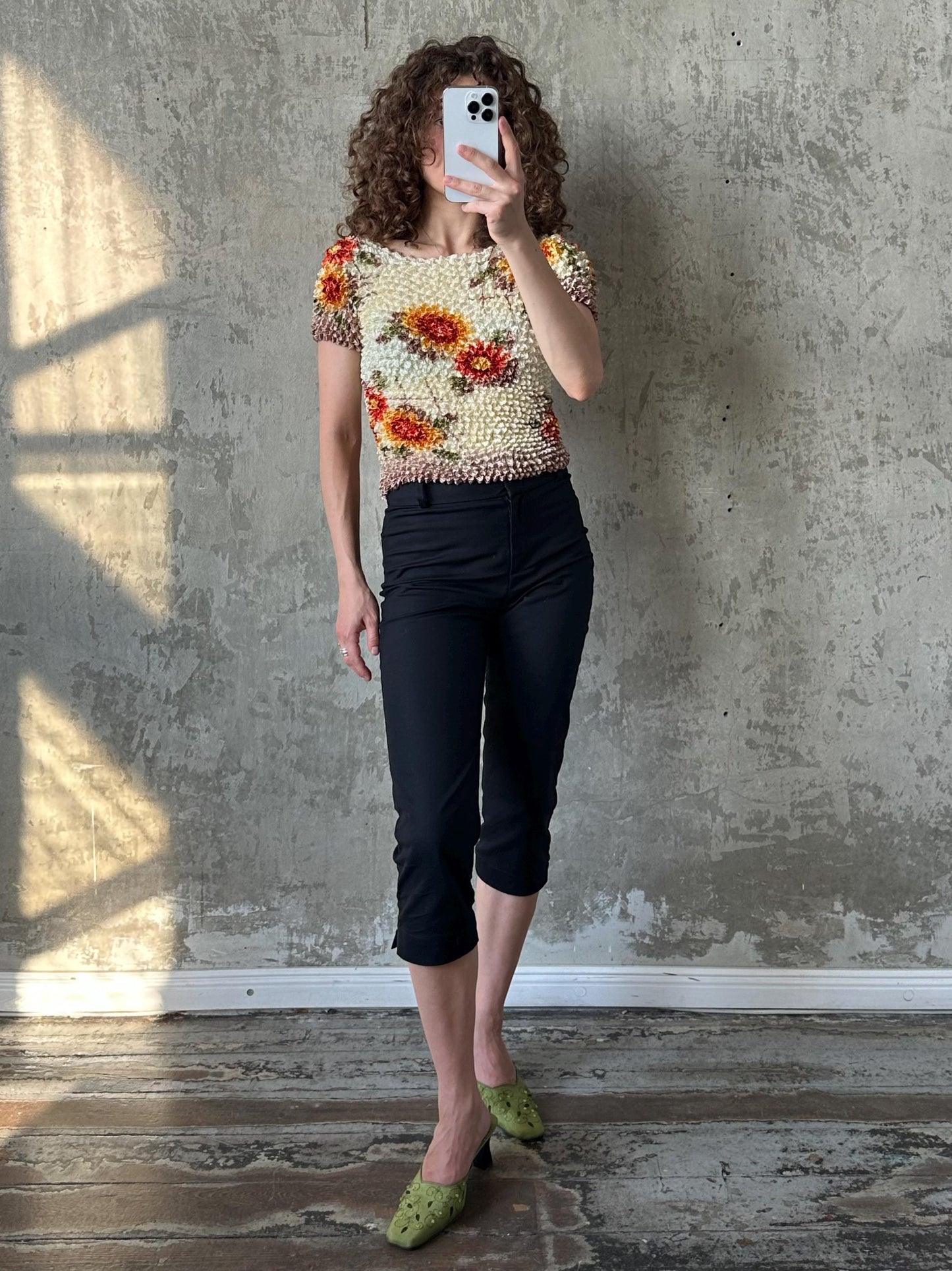 A woman with curly hair is taking a selfie with a white phone, wearing a floral top and black pants, and green shoes.