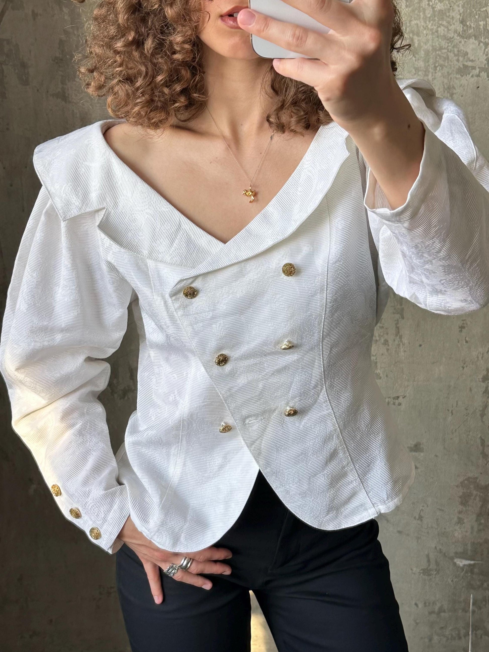 A woman with curly hair is taking a selfie with a white blouse and black pants.