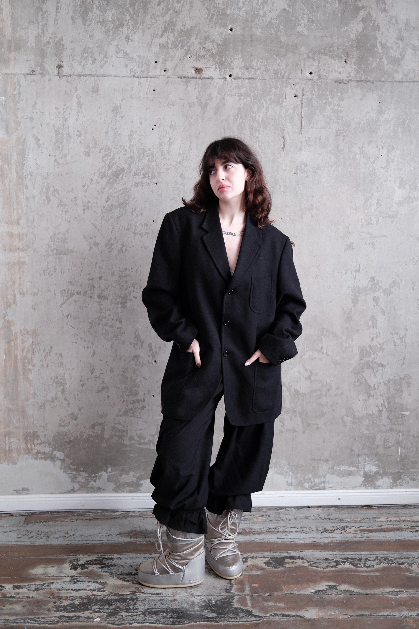 A person wearing a black suit stands in front of a concrete wall, with their head turned to the side.