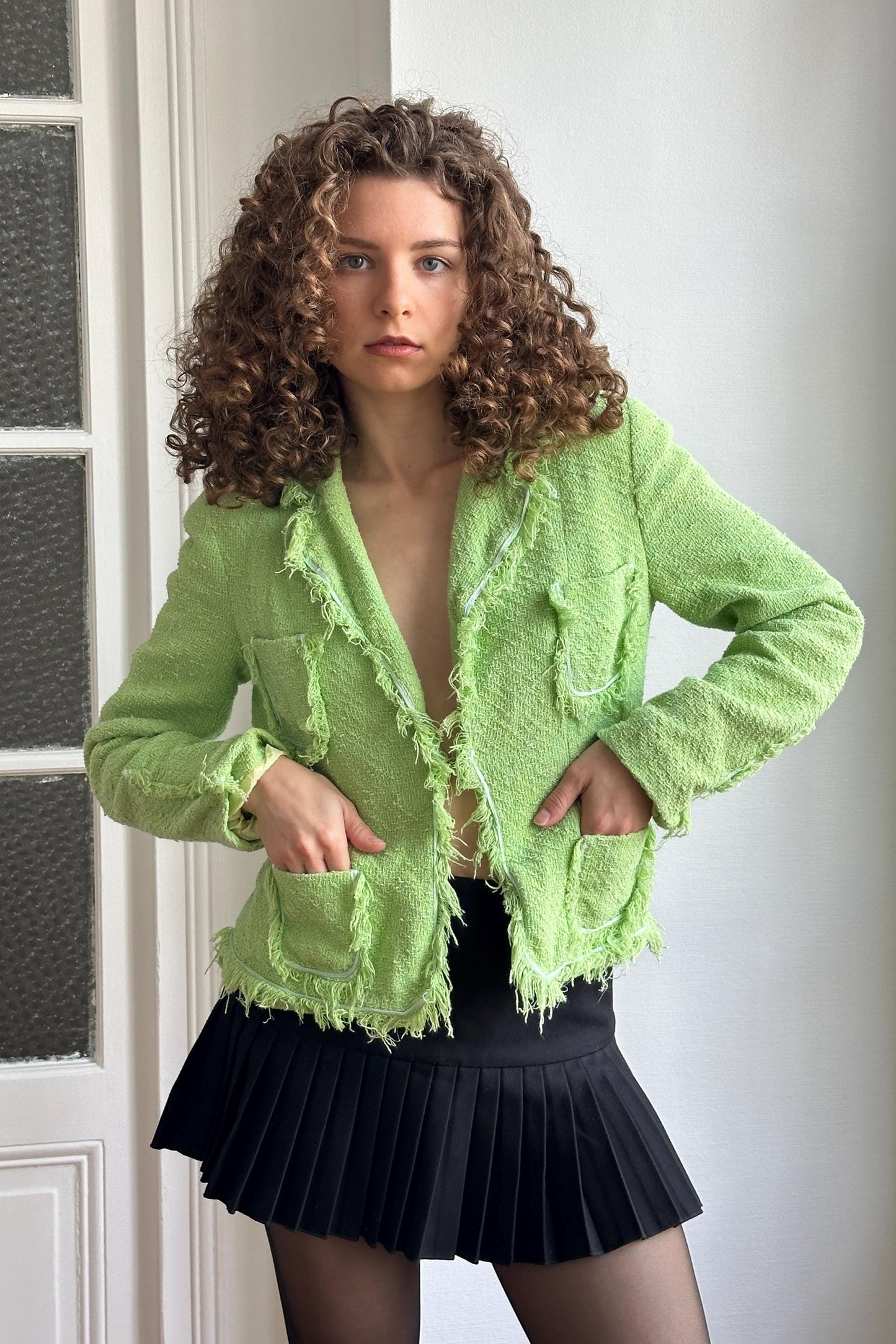 A young woman with curly hair wearing a lime green jacket and a black pleated skirt stands in front of a white door.