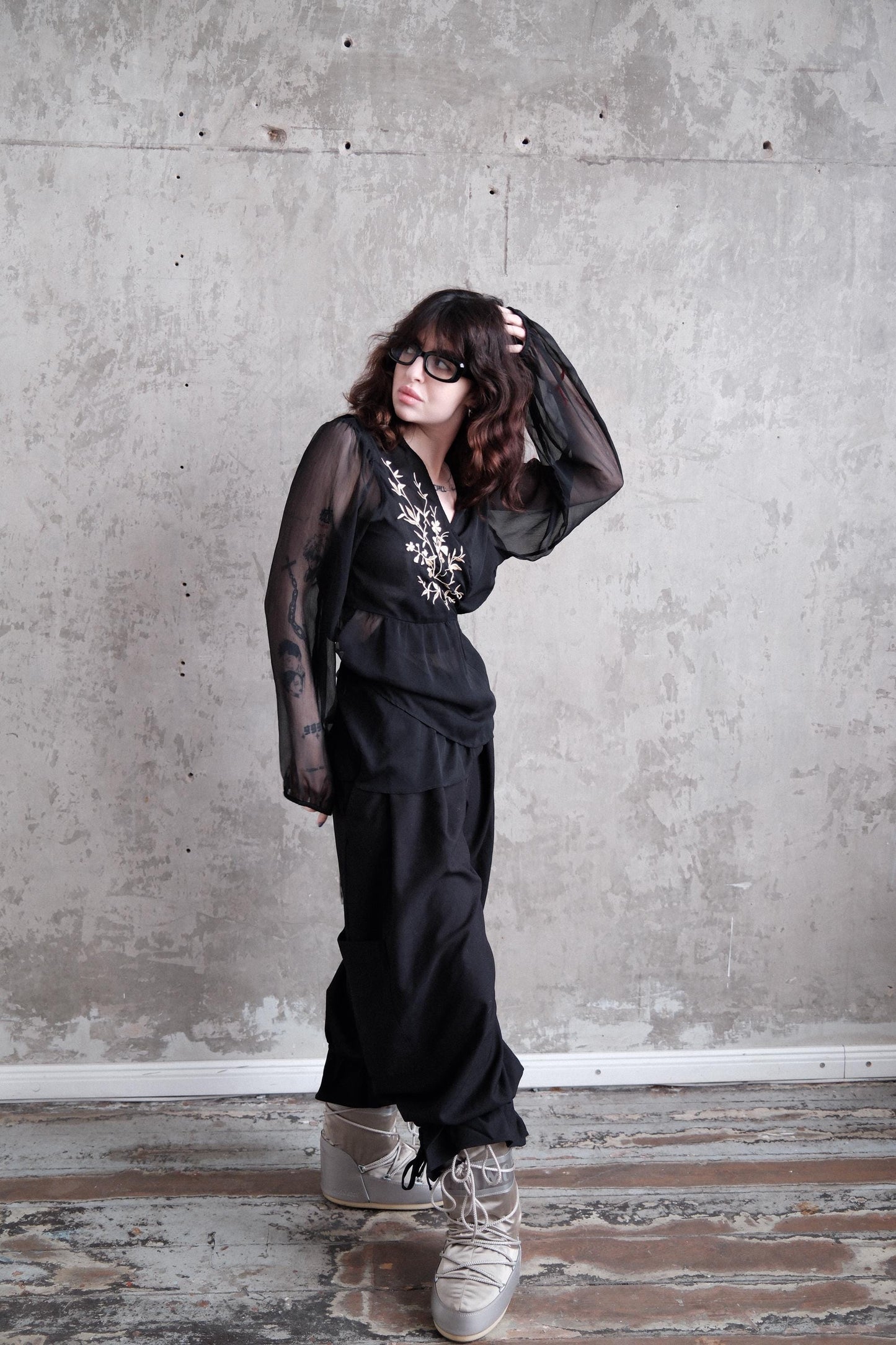 A woman in a black outfit with sheer sleeves stands in a room with a concrete wall.