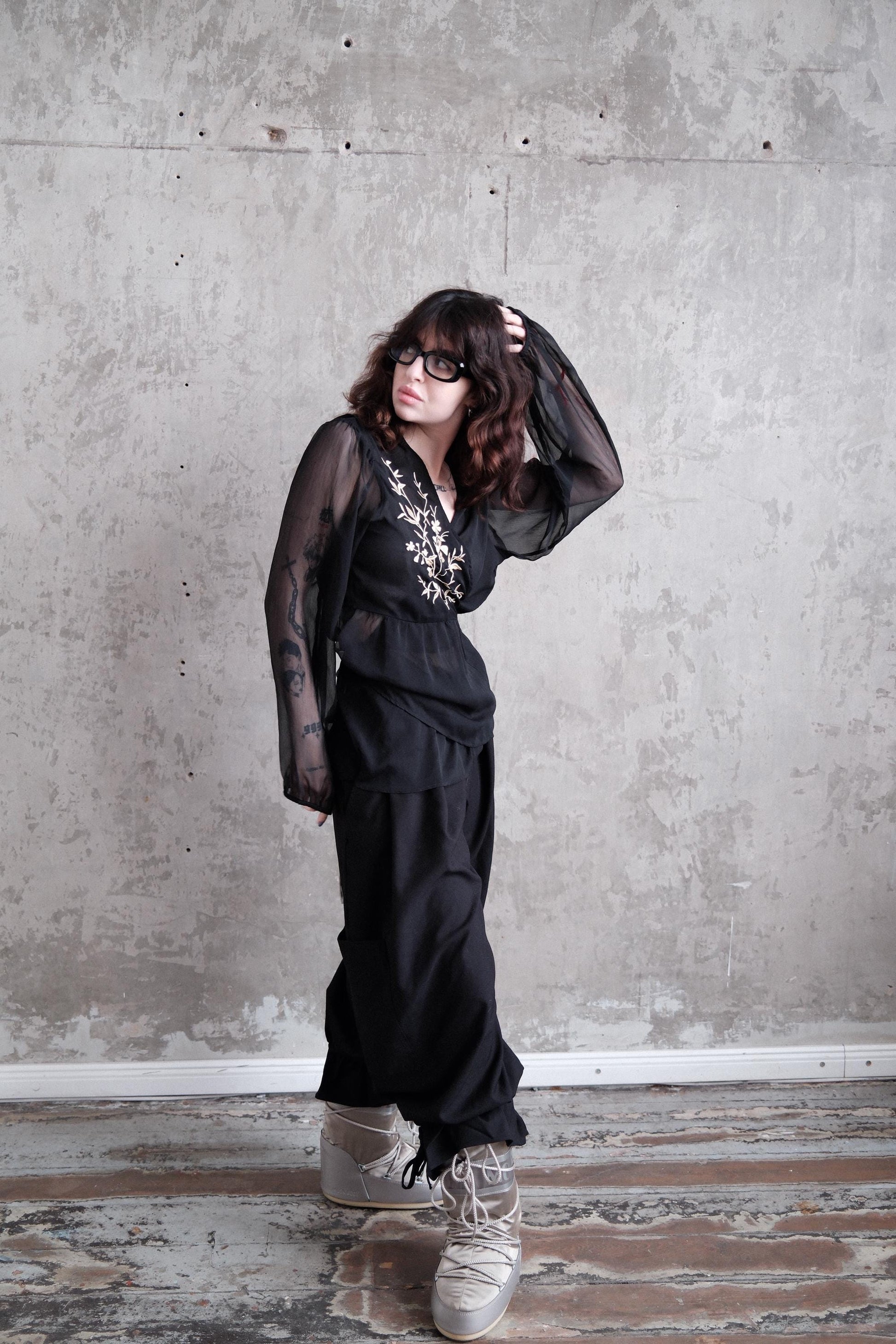A woman in a black outfit with sheer sleeves stands in a room with a concrete wall.