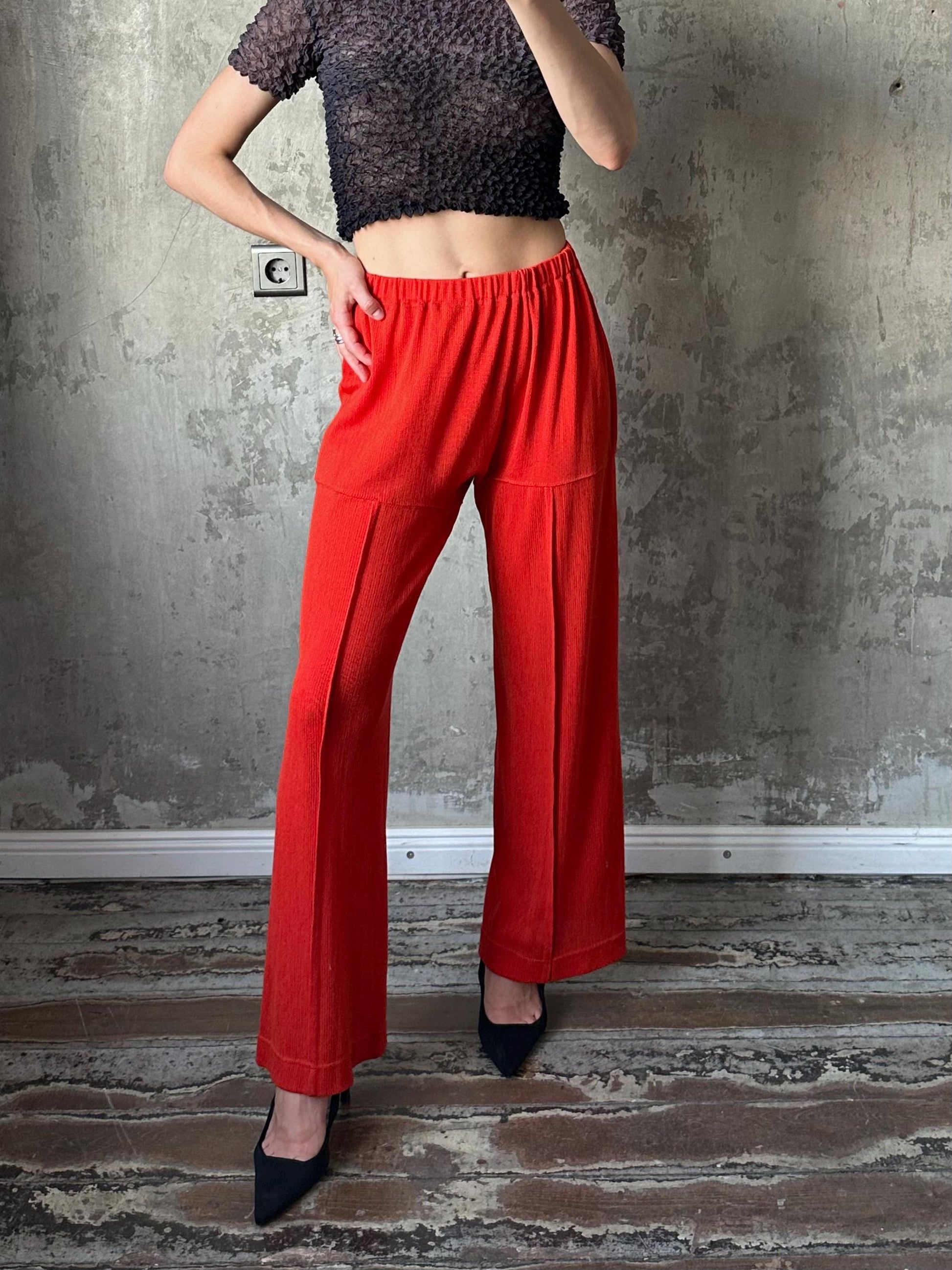 A person wearing a black crop top and red wide-leg pants is standing in front of a concrete wall.