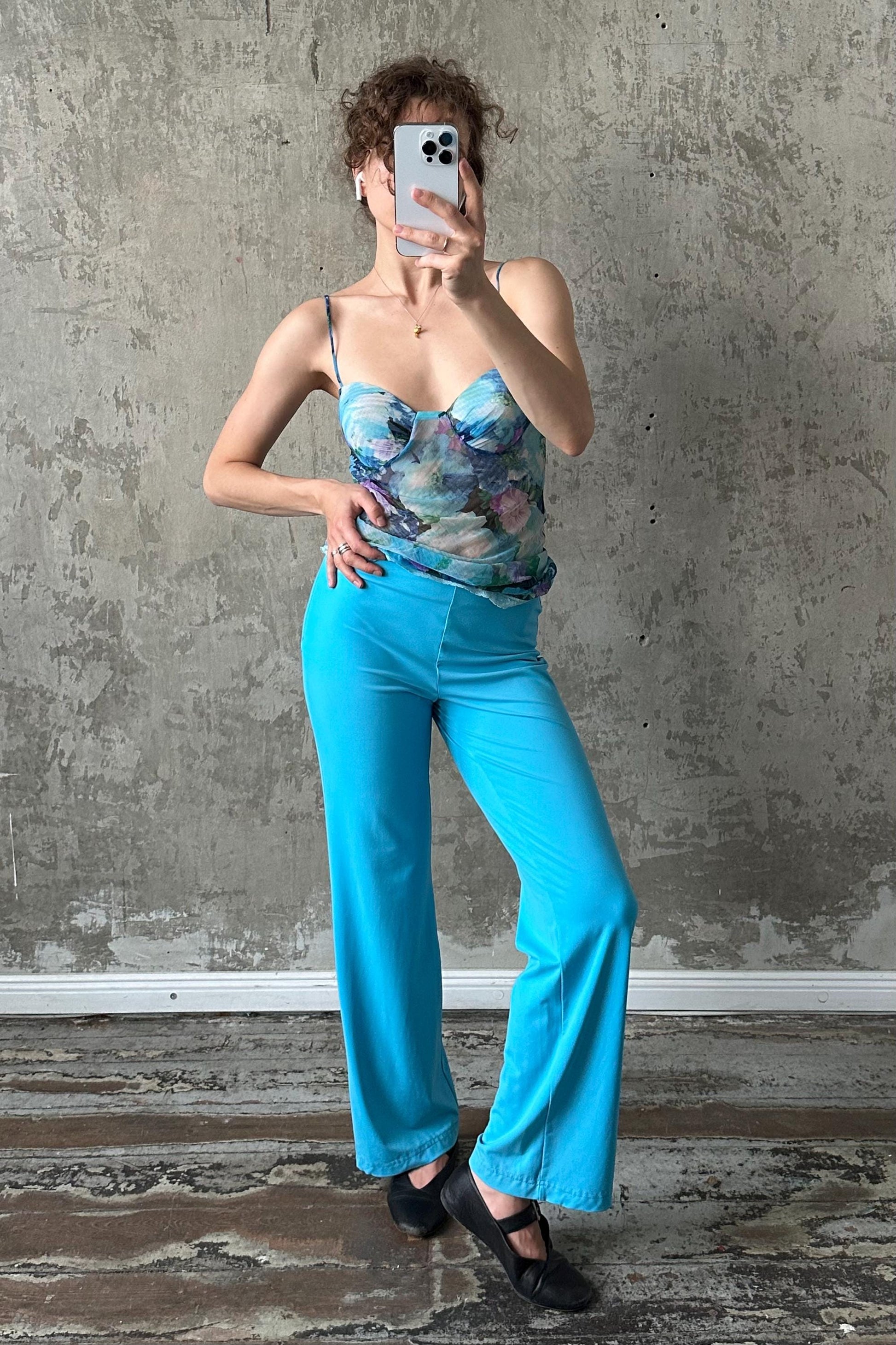 A woman is taking a selfie with her phone, wearing a blue floral top and blue wide-leg pants, standing in front of a concrete wall.