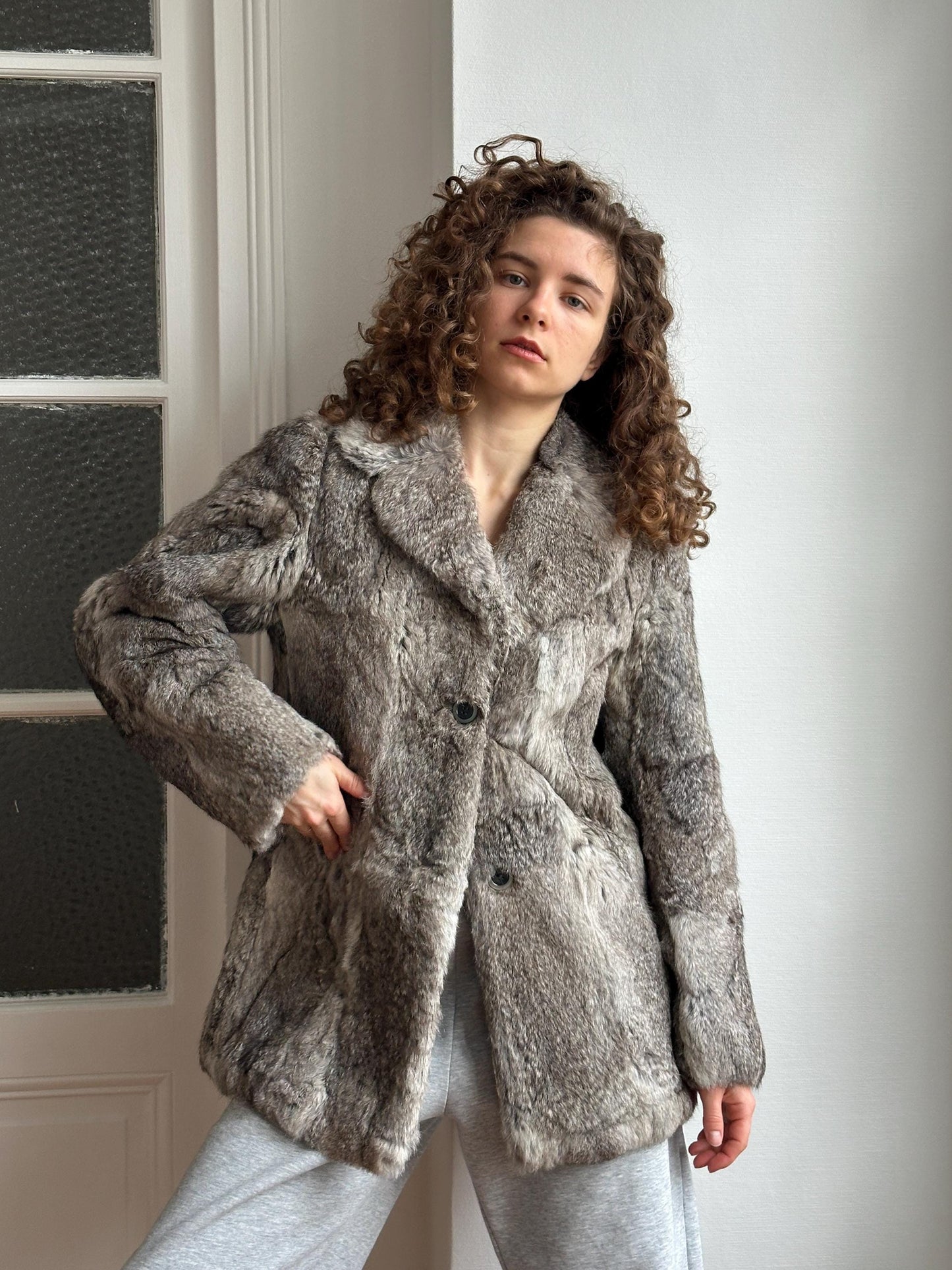 A woman with curly hair wearing a gray fur coat stands in front of a white wall.