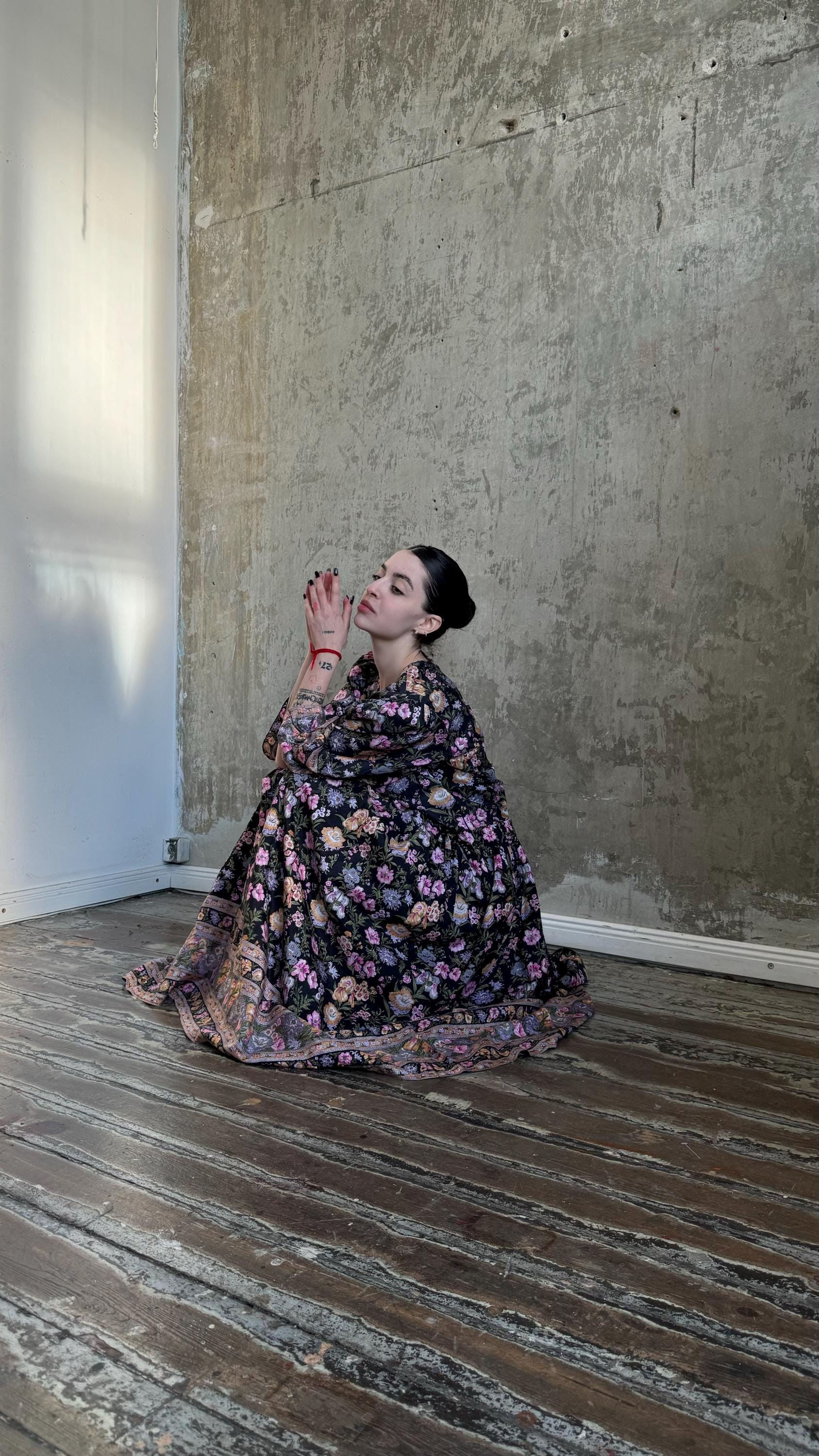A woman in a floral dress kneels on a wooden floor, her hands clasped together in a prayer position.