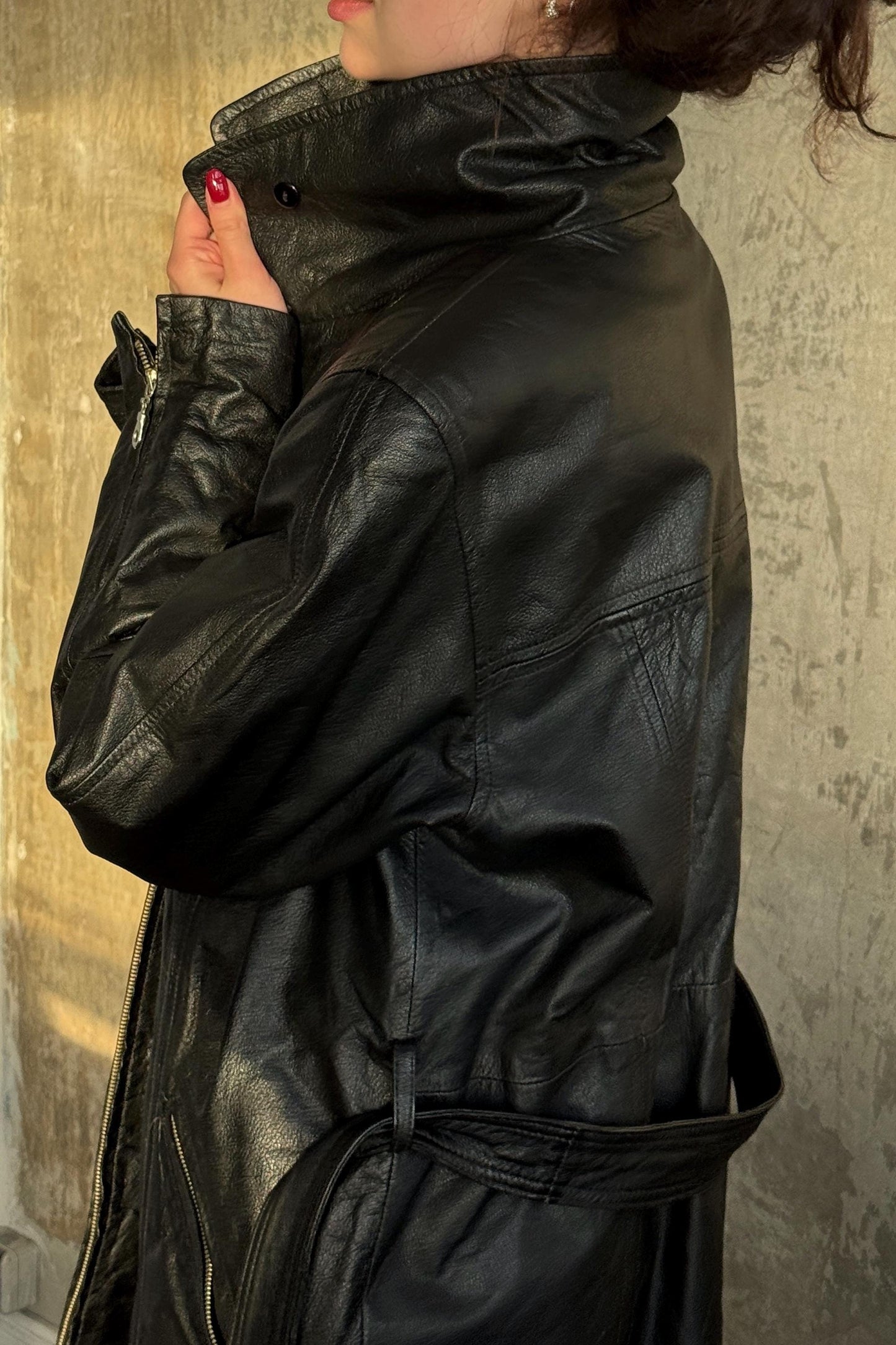 A person wearing a black leather jacket stands against a concrete wall.