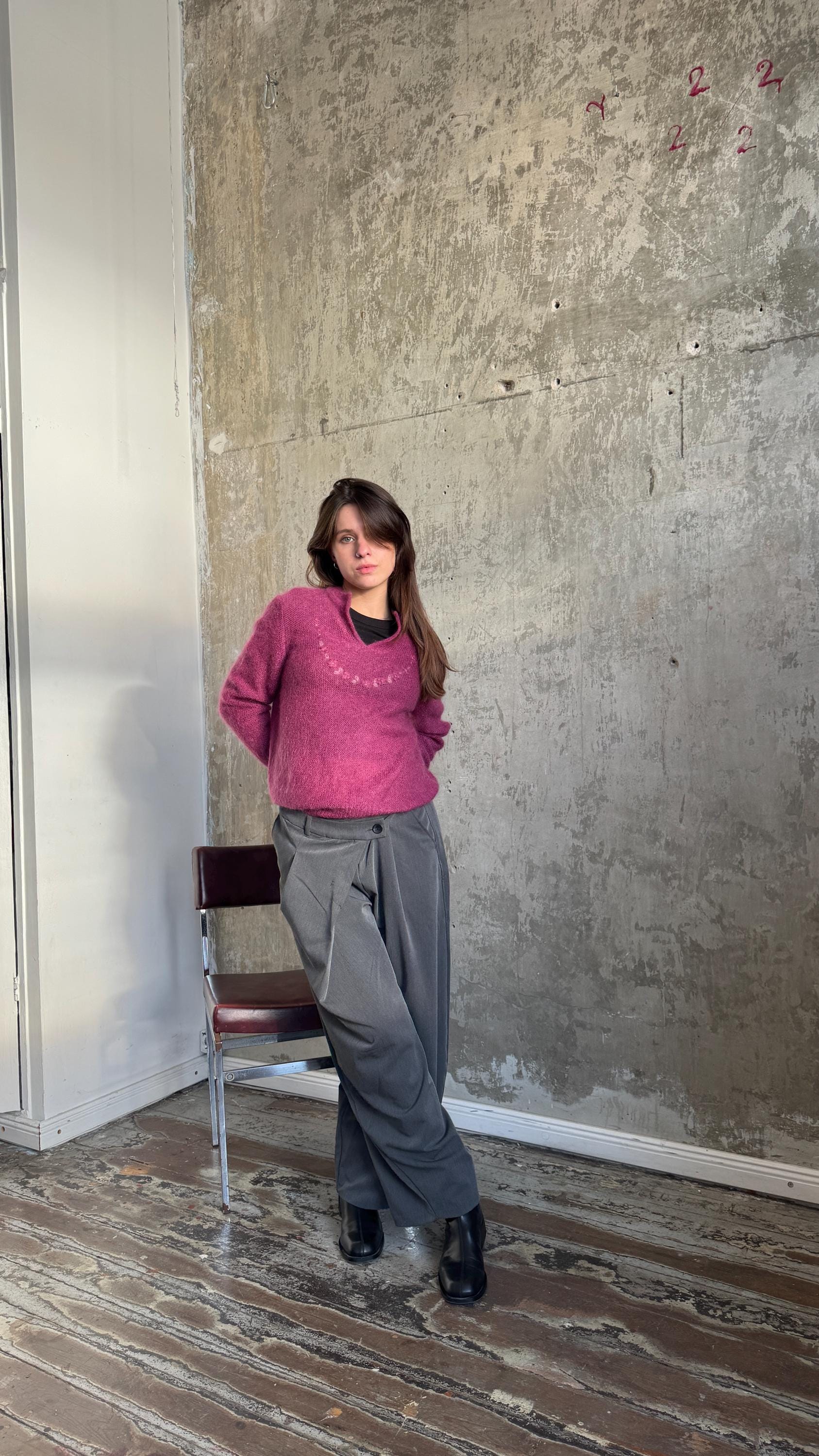 A woman in a pink sweater and gray pants stands in a room with a wooden floor and a concrete wall.