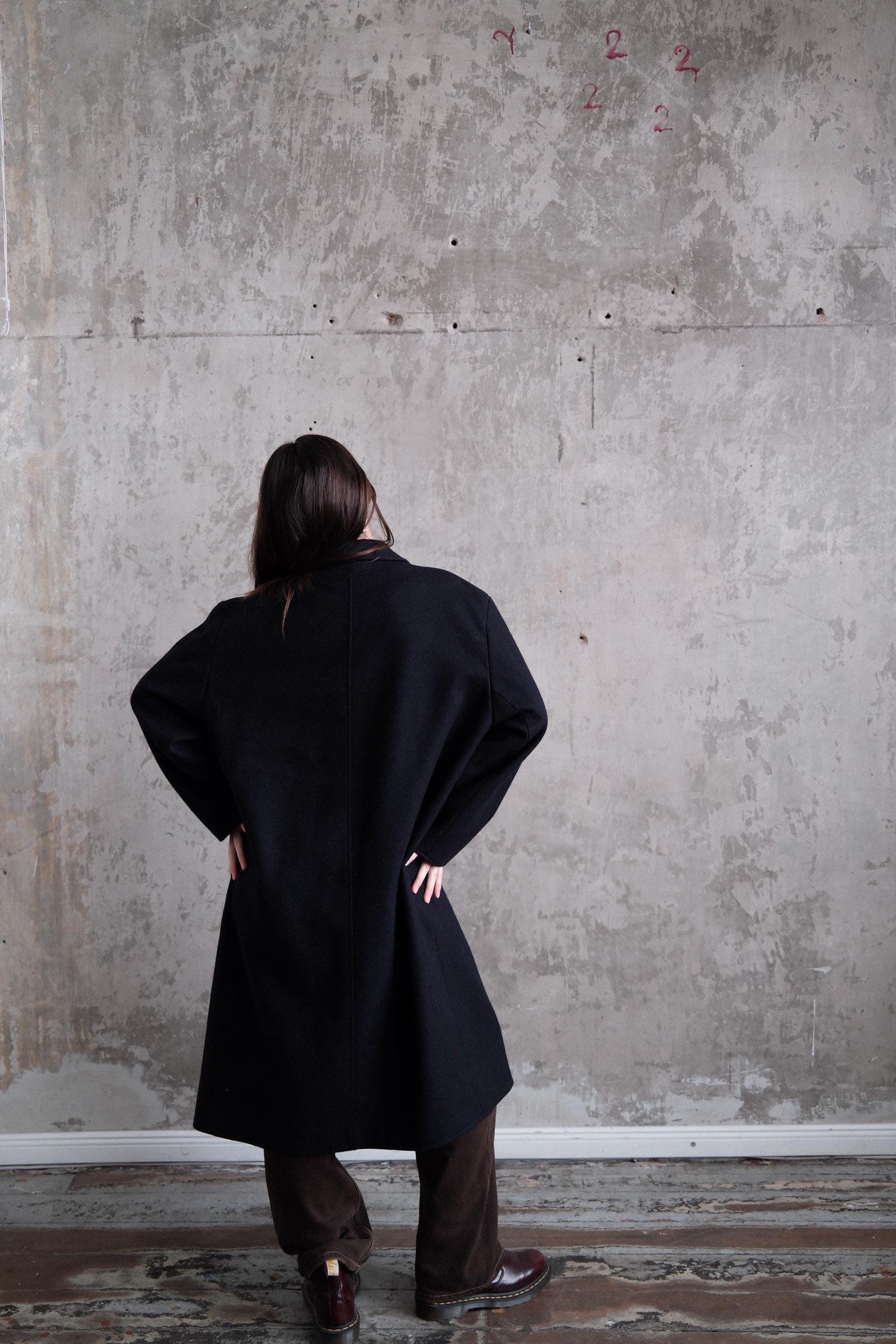 A person wearing a black coat stands in a room with a concrete wall and a red graffiti mark.