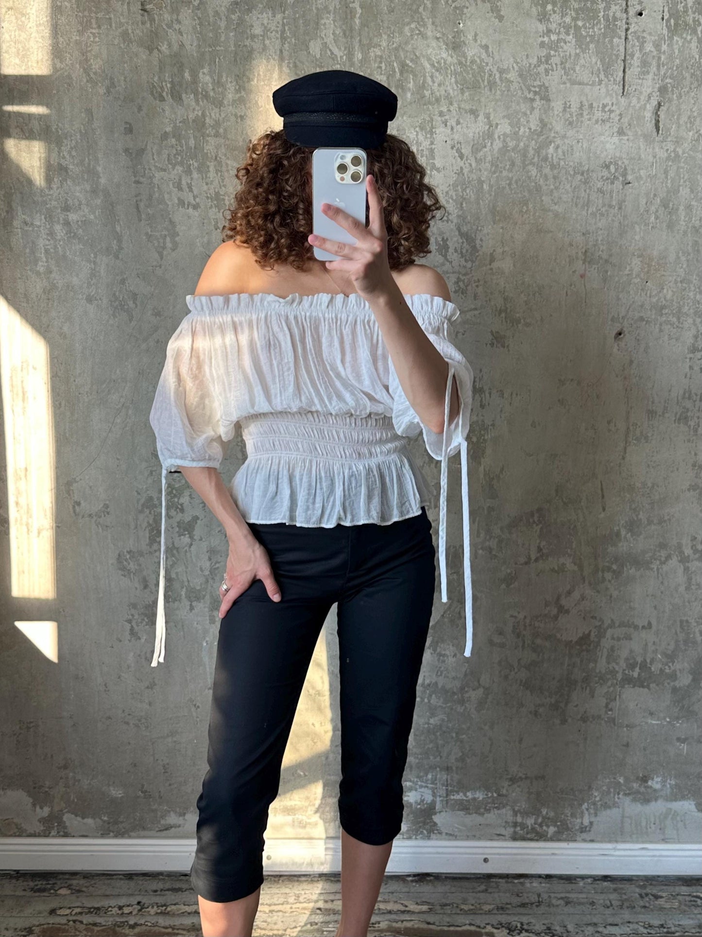 A person is taking a selfie with a smartphone, wearing a white off-the-shoulder top and black pants.