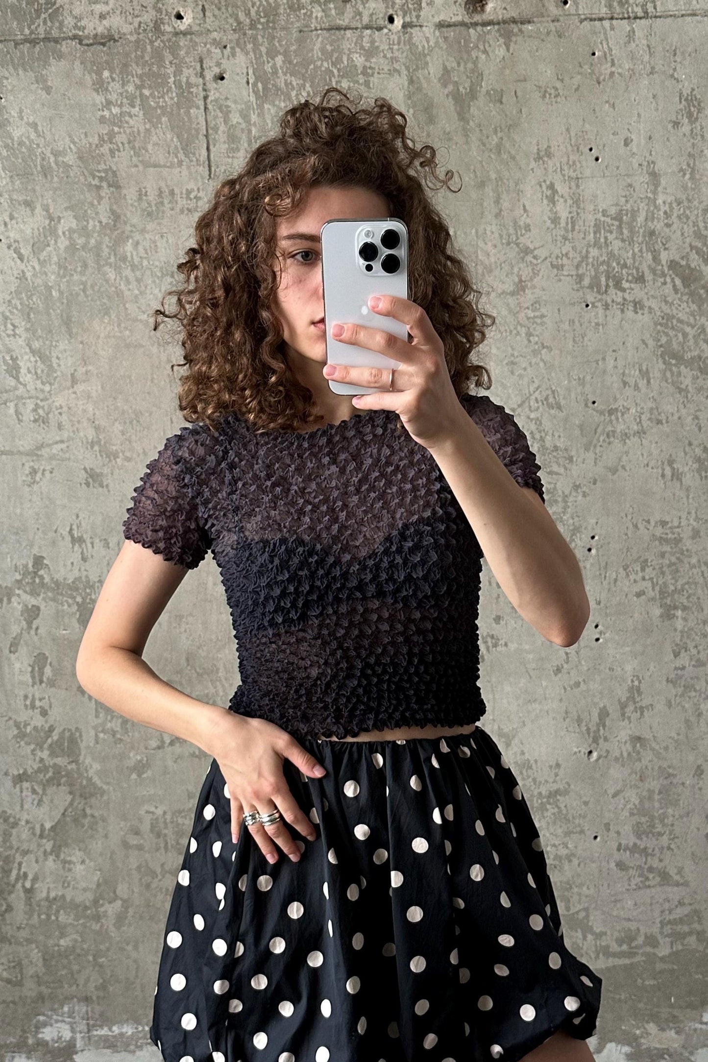 A woman with curly hair is taking a selfie with her iPhone, wearing a black top with a polka dot pattern and a black and white polka dot skirt.