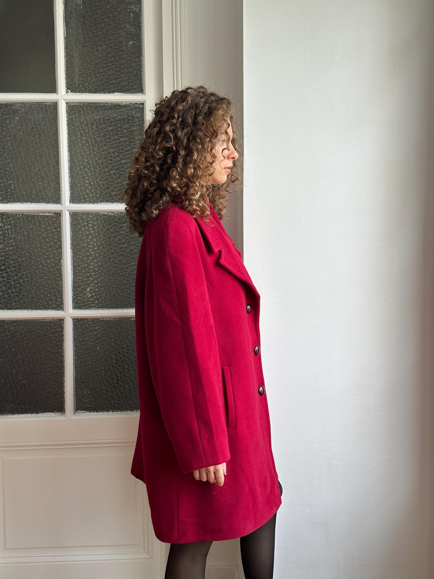 A person wearing a red coat is floating in mid-air, with their head tilted back and arms outstretched, against a plain white wall.