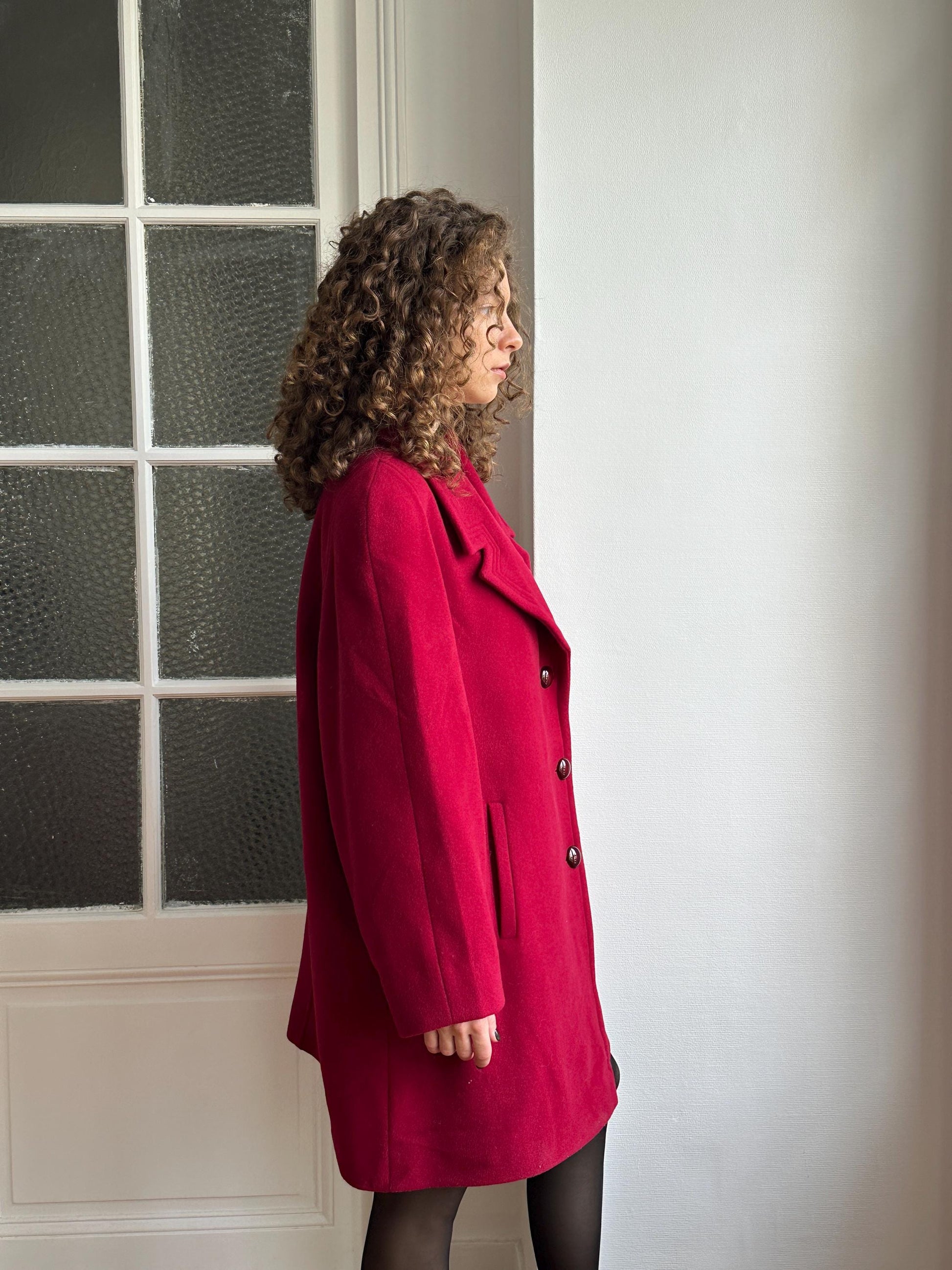 A person wearing a red coat is floating in mid-air, with their head tilted back and arms outstretched, against a plain white wall.