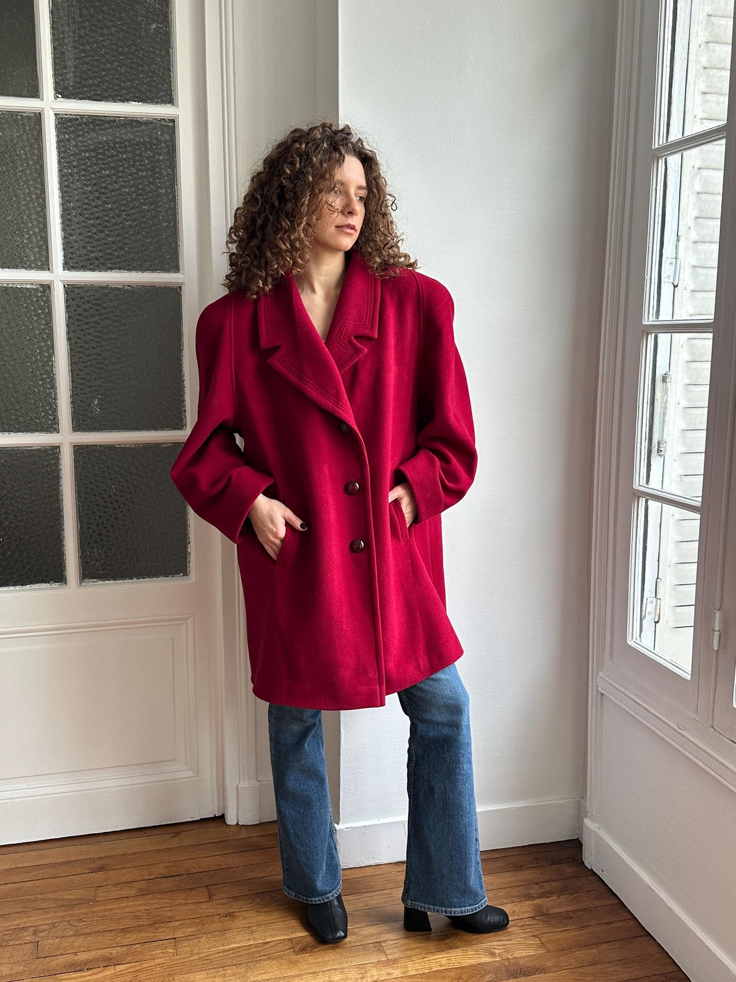 A person wearing a red coat and blue jeans stands in a room with a window.