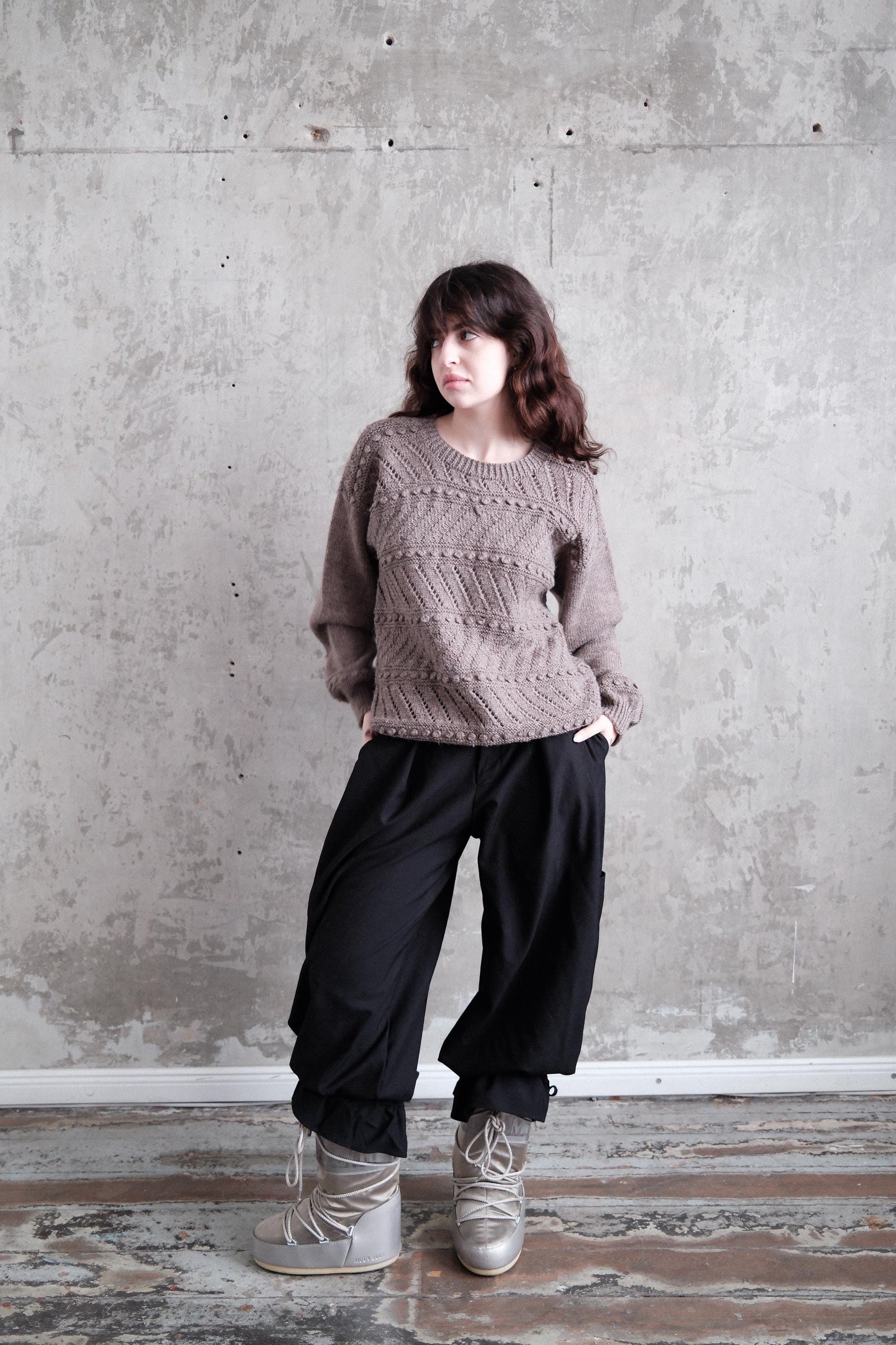 A person wearing a gray sweater and black pants stands against a concrete wall, with their head tilted back and hands in their pockets.