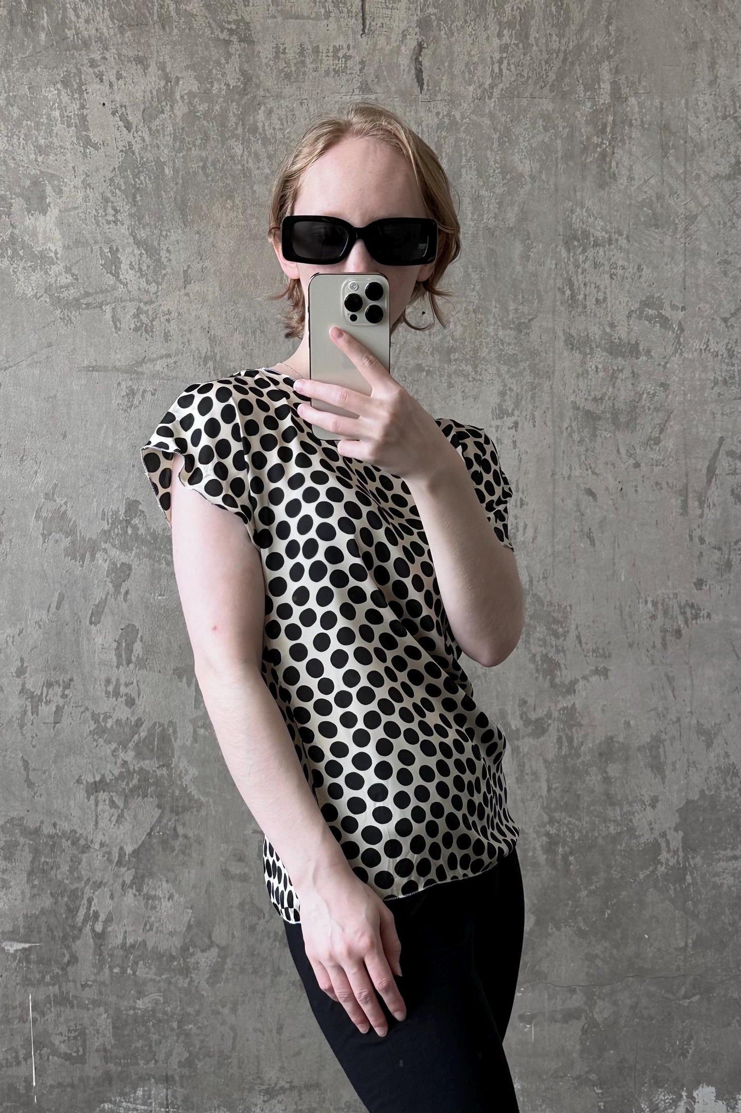 A person is taking a selfie with a smartphone, wearing a black and white polka dot blouse and sunglasses.
