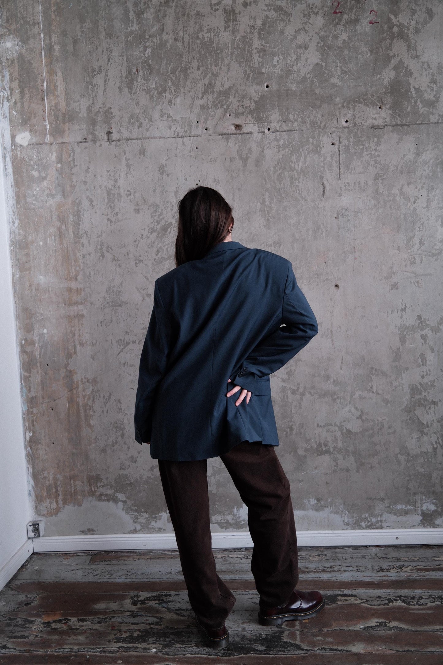 A person wearing a dark jacket and brown pants is standing in front of a concrete wall, with their back turned to the camera.
