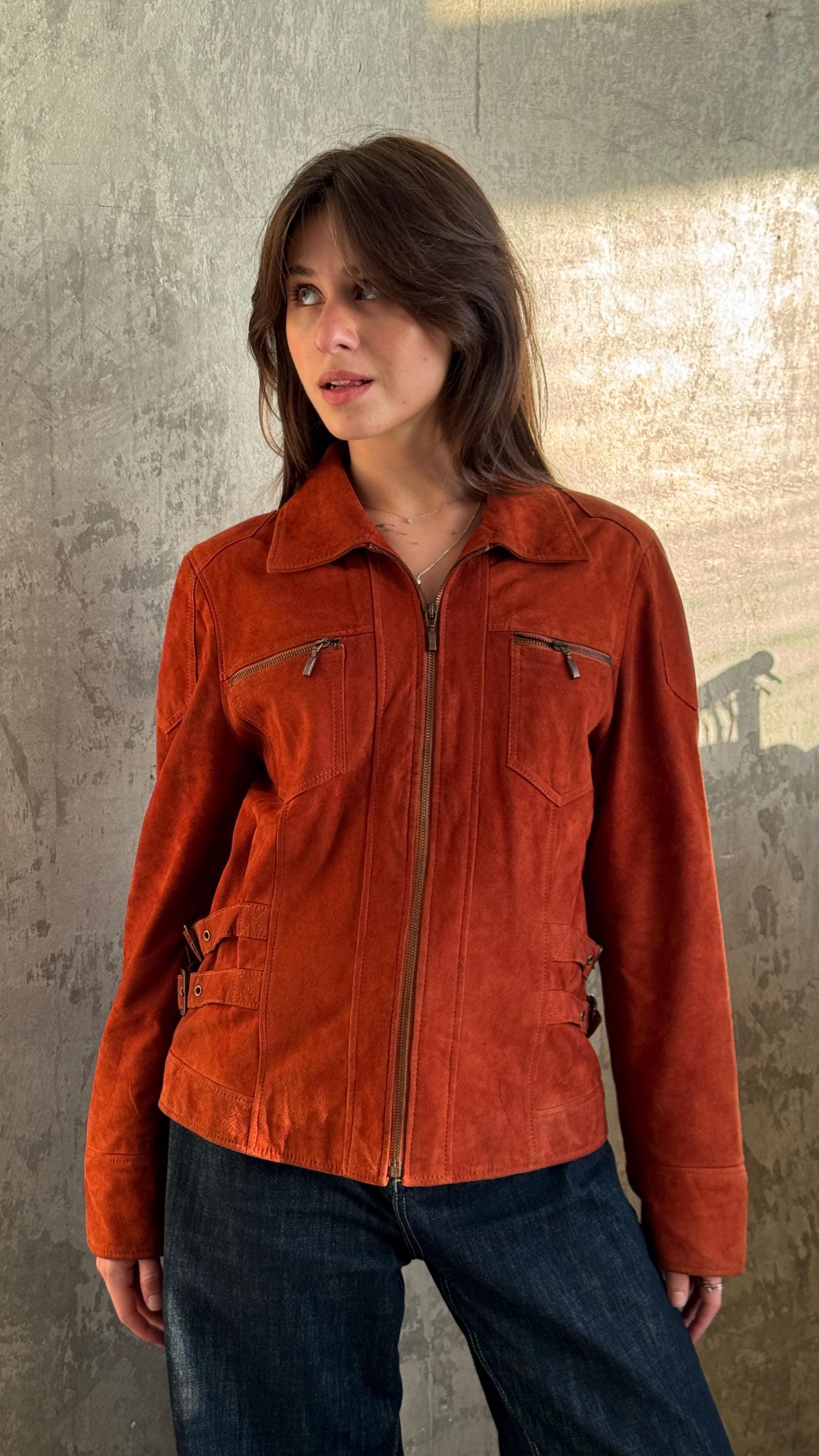 A woman wearing a red suede jacket stands in front of a concrete wall, looking off to the side.