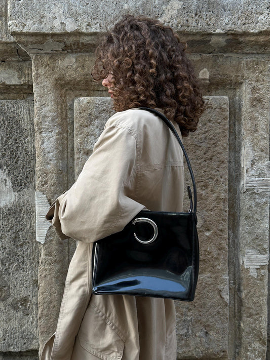 A woman with curly hair is standing in front of a stone wall, wearing a beige coat and carrying a black purse with a silver circular logo.