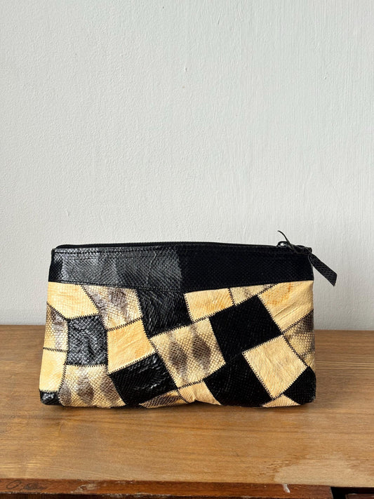 A black and beige patterned clutch purse is displayed on a wooden surface against a white wall.