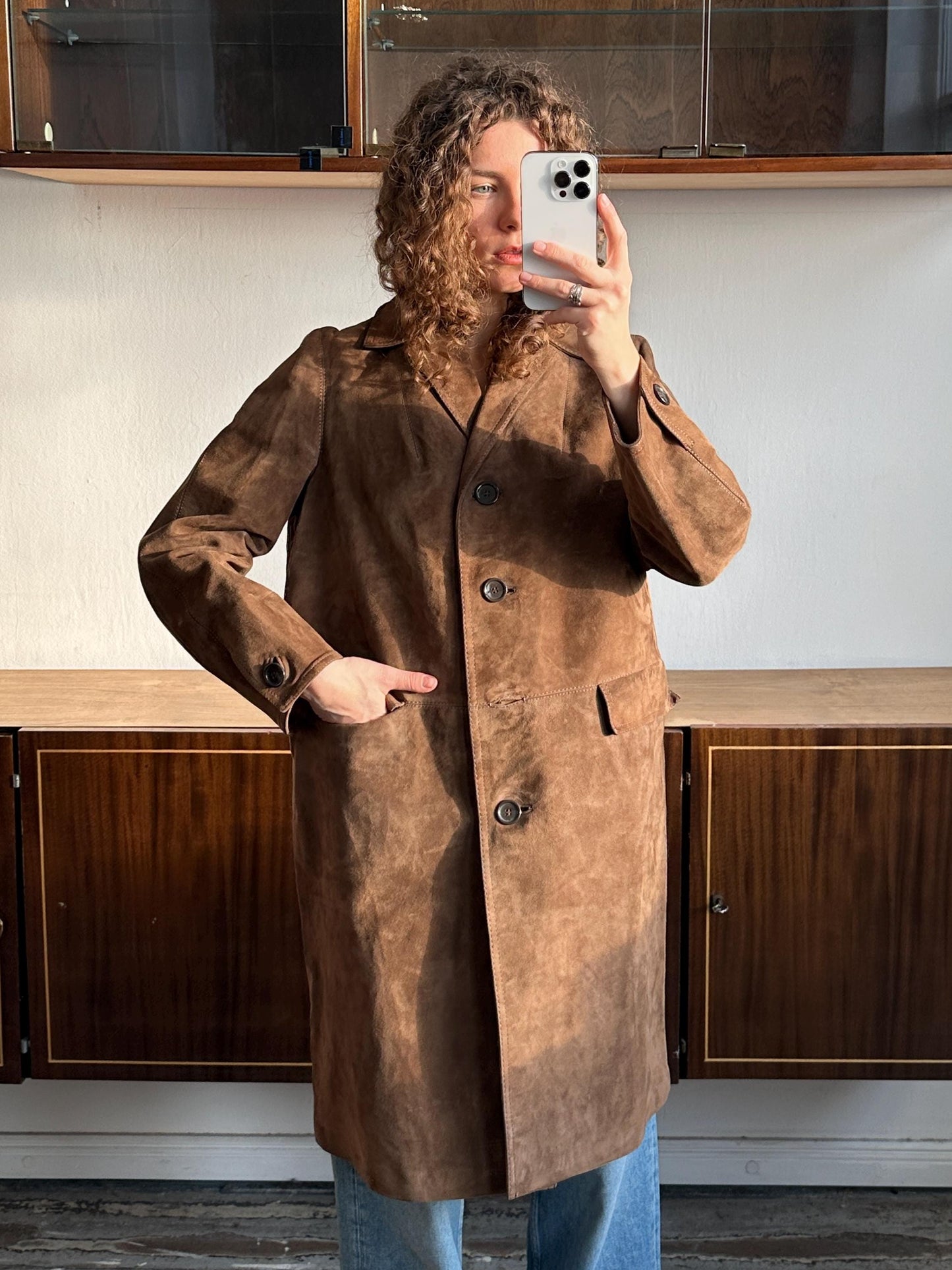 A woman wearing a brown suede coat is taking a selfie with her iPhone in front of a wooden cabinet.