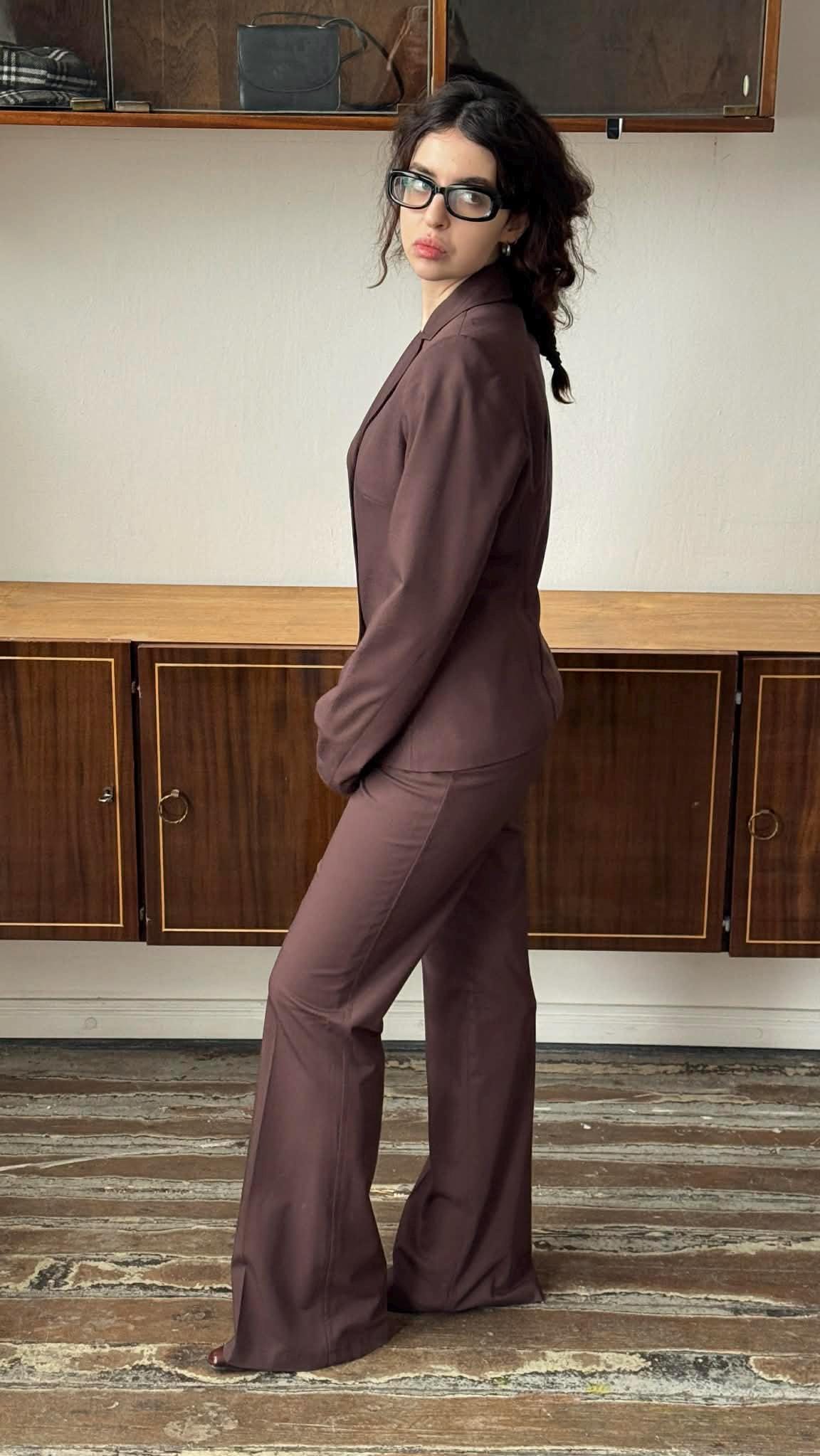 A woman wearing a brown suit stands in a room with wooden cabinets in the background.