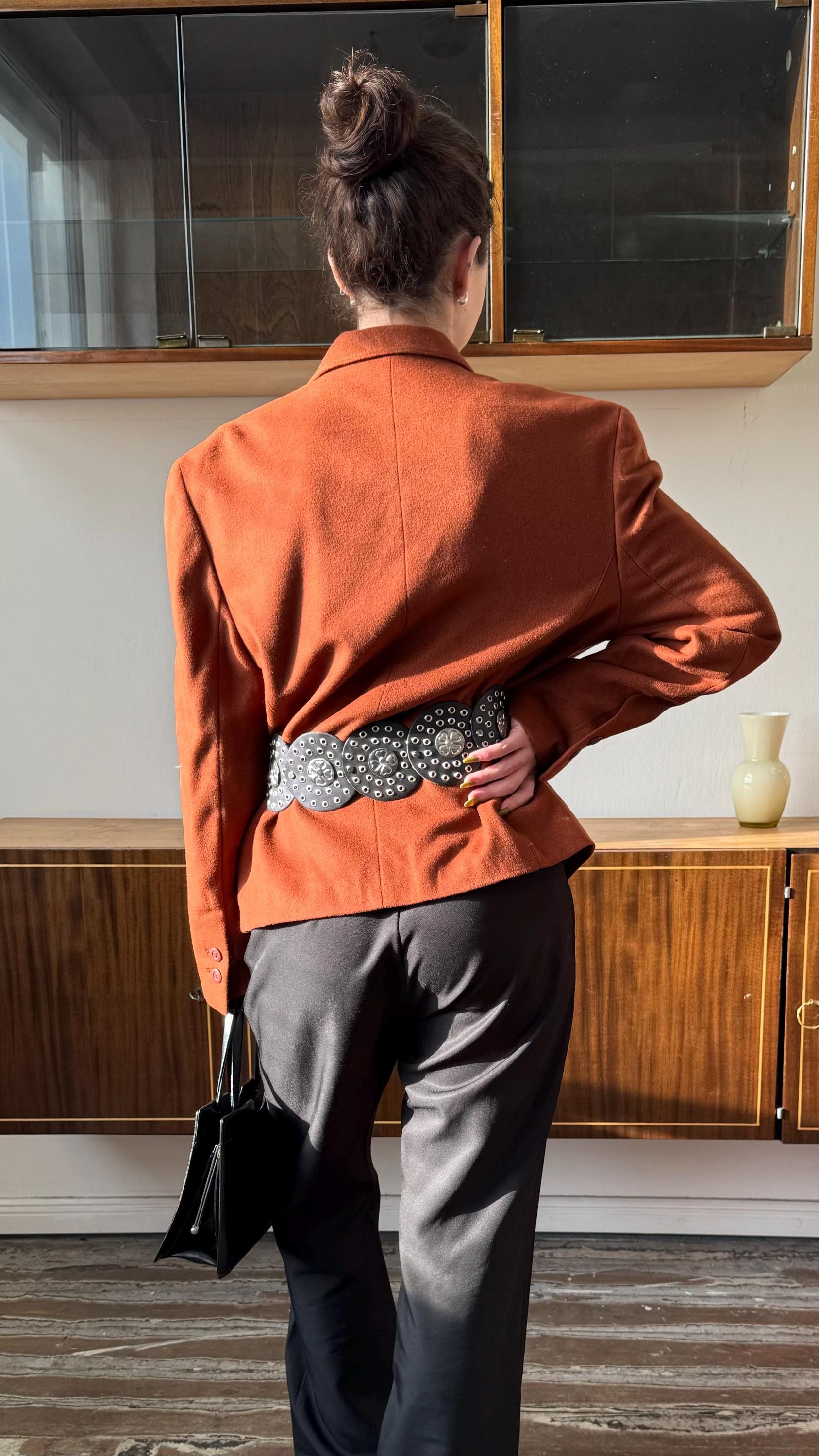 A woman wearing a red blazer and holding a black purse stands in front of a wooden cabinet.