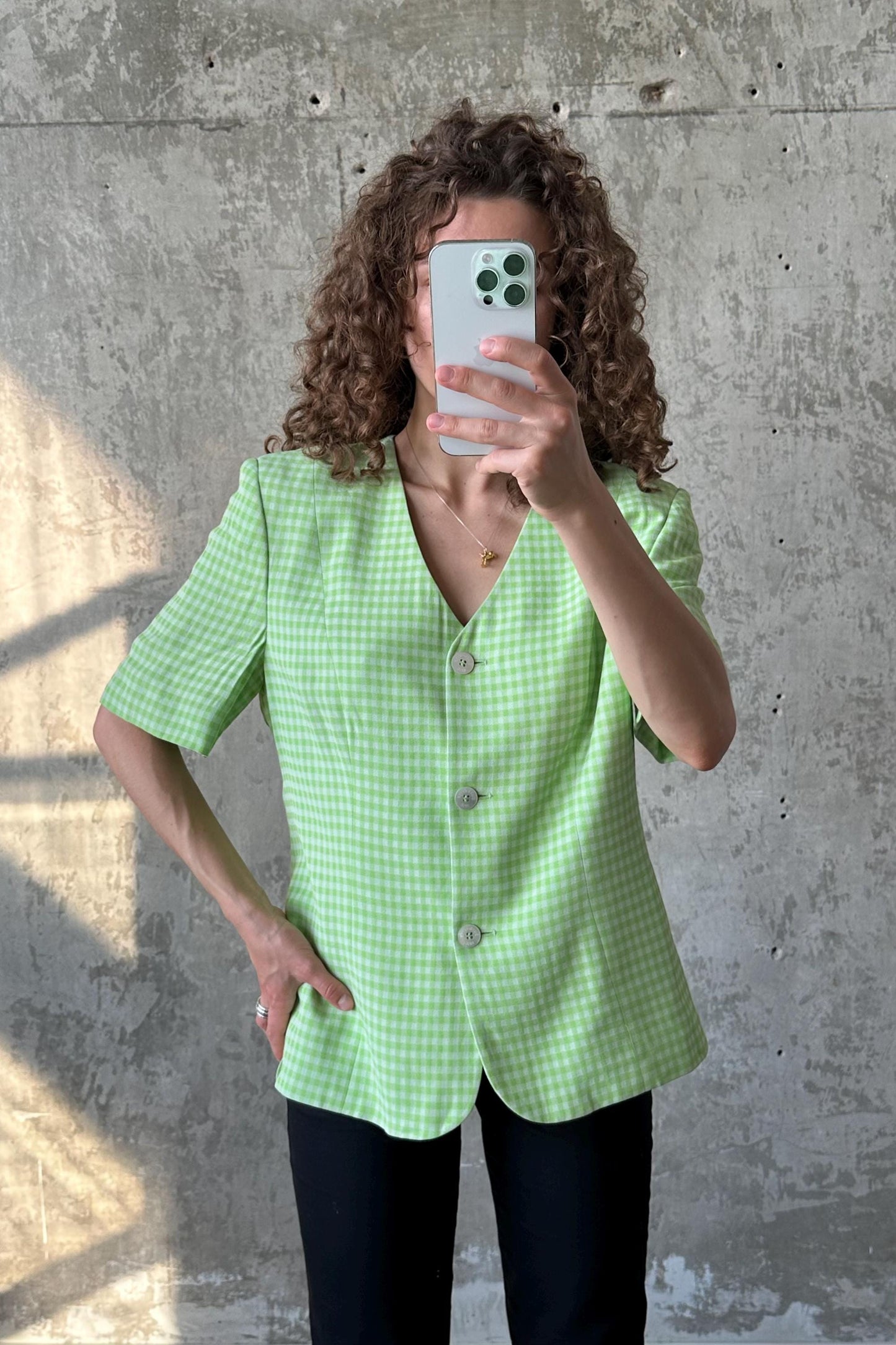 A woman with curly hair is taking a selfie with her phone, wearing a green and white checkered blazer.