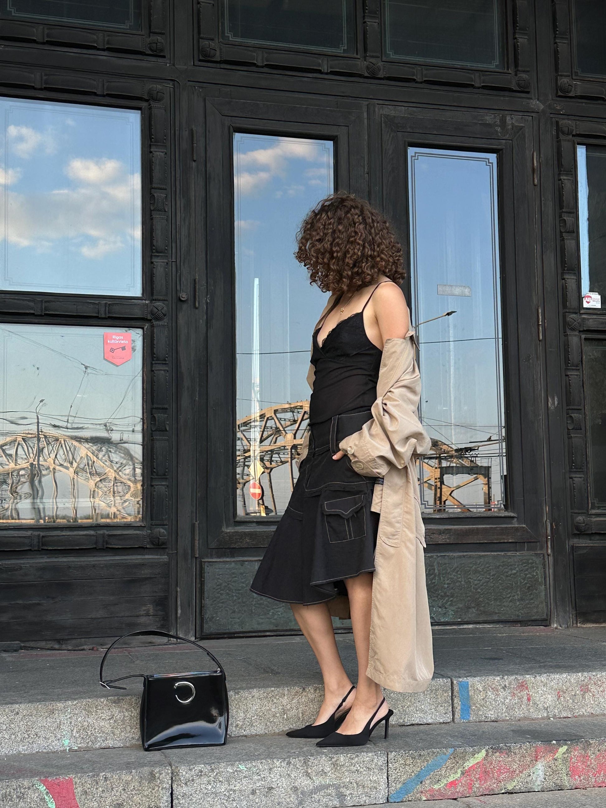 A woman in a black dress and beige coat stands on a city street, with a black purse and a black handbag on the ground in front of her.