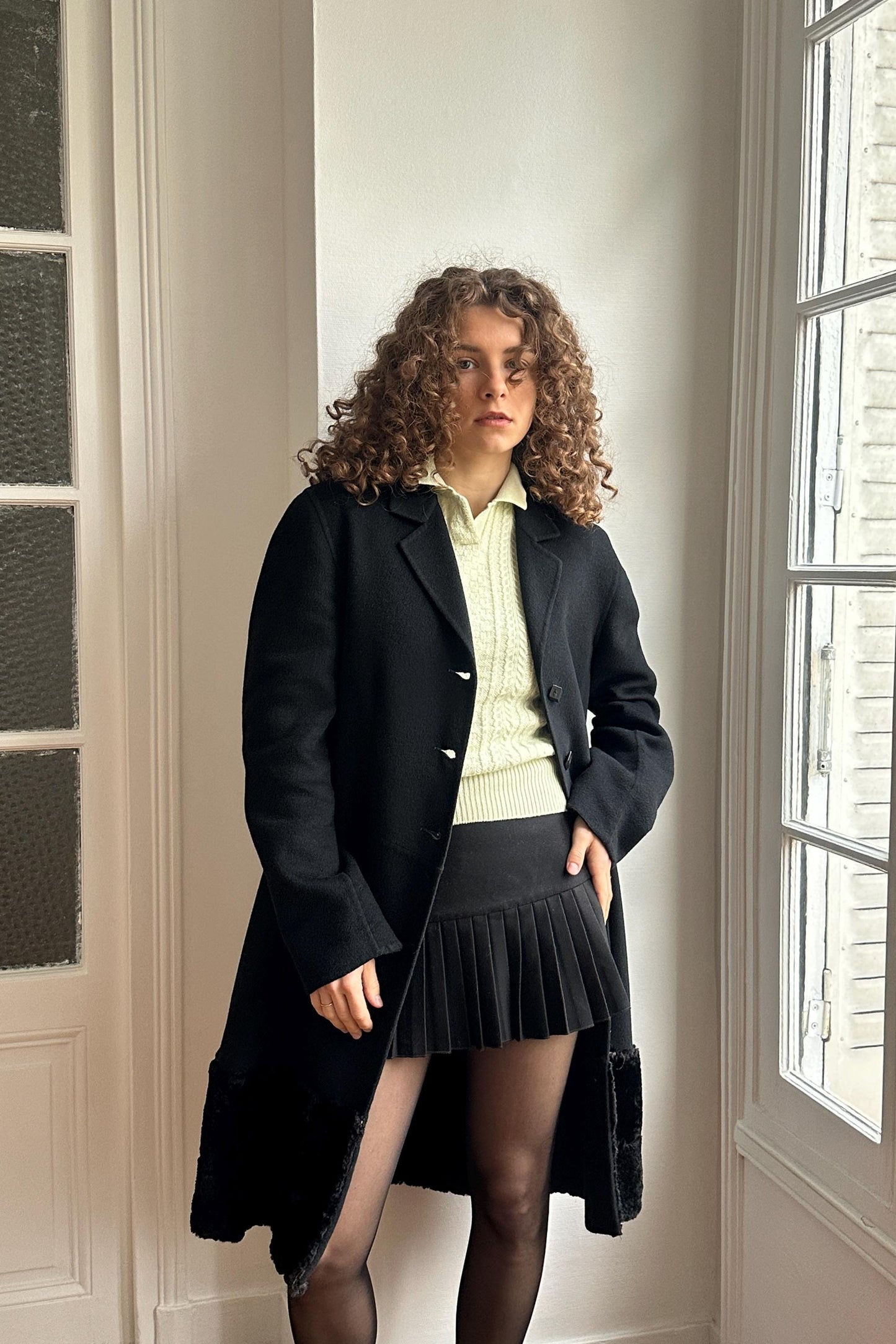 A woman with curly hair wearing a black coat, white shirt, and black skirt stands in a room with a window.