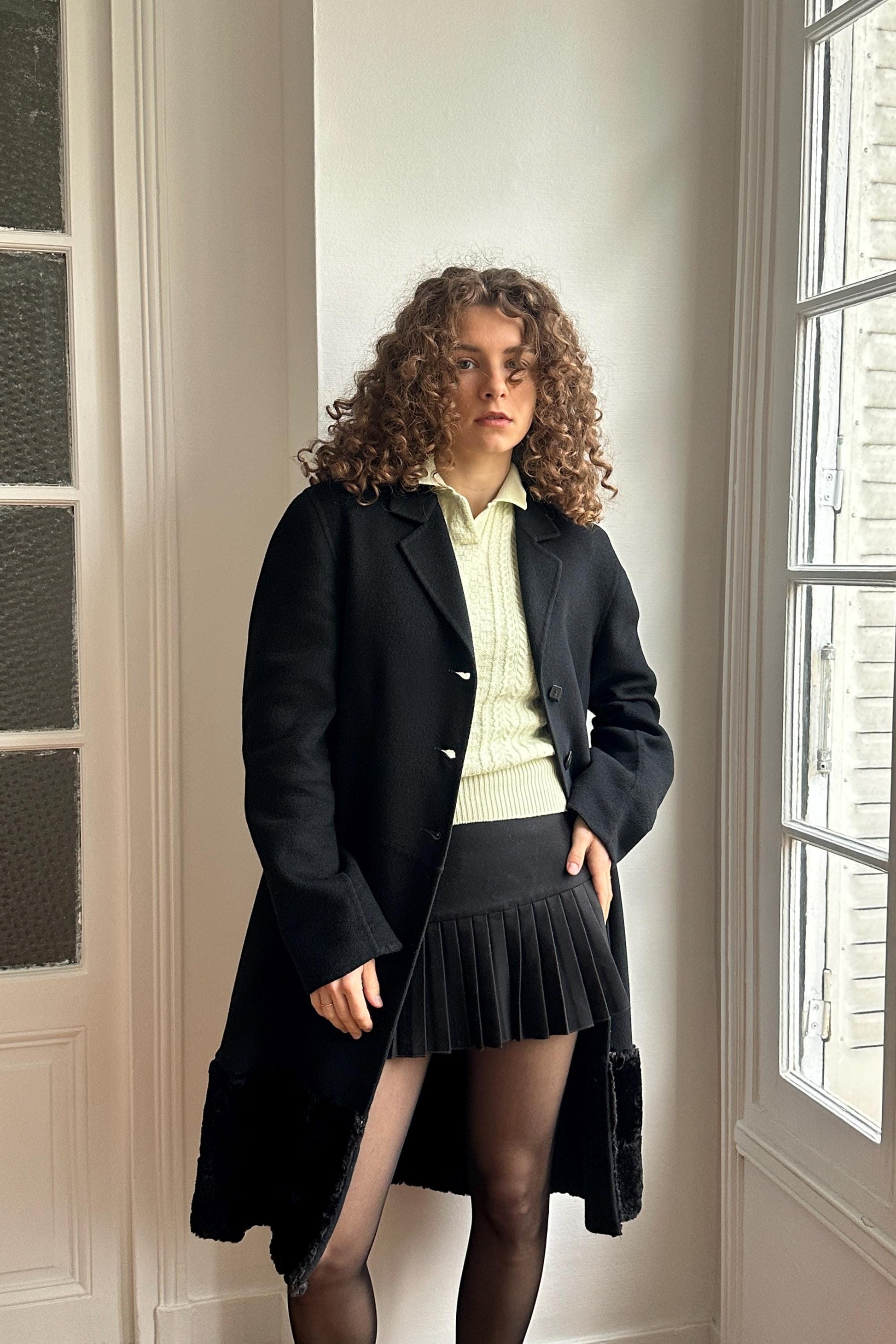 A woman with curly hair wearing a black coat, white shirt, and black skirt stands in a room with a window.