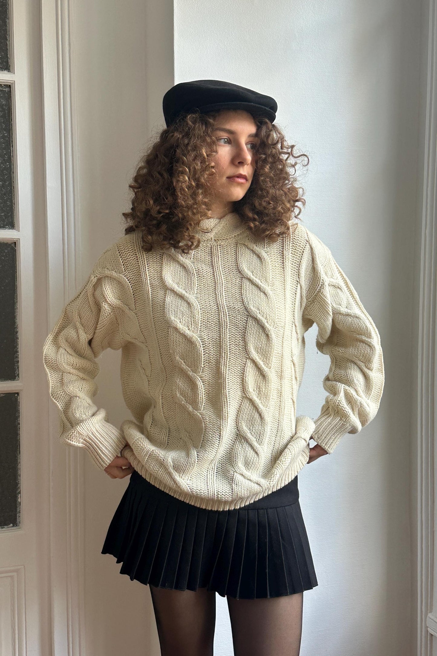A young woman with curly hair stands in front of a white wall, wearing a beige sweater, a black skirt, and a black hat.