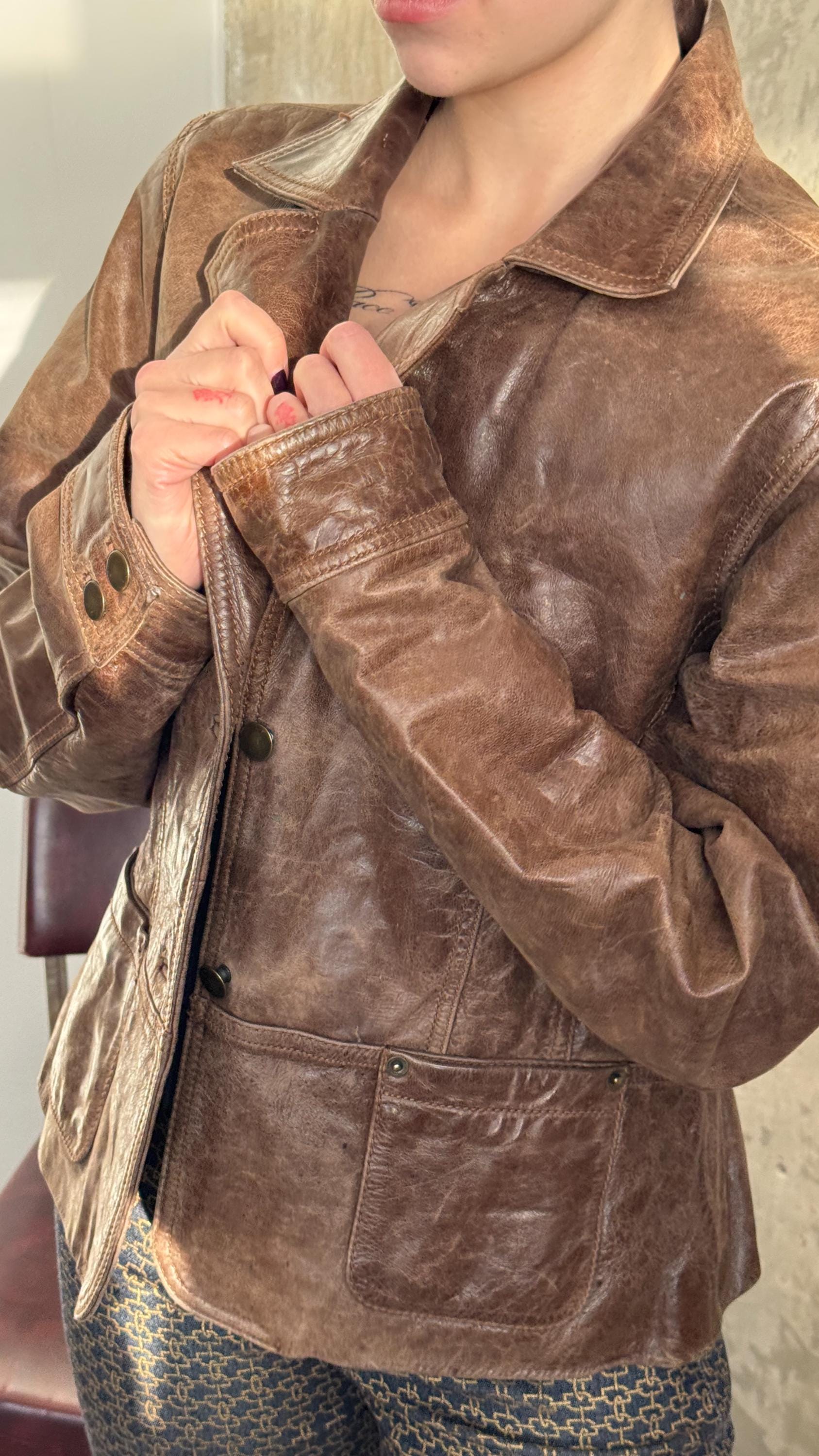 A person wearing a brown leather jacket and patterned pants, with their hands clasped together.