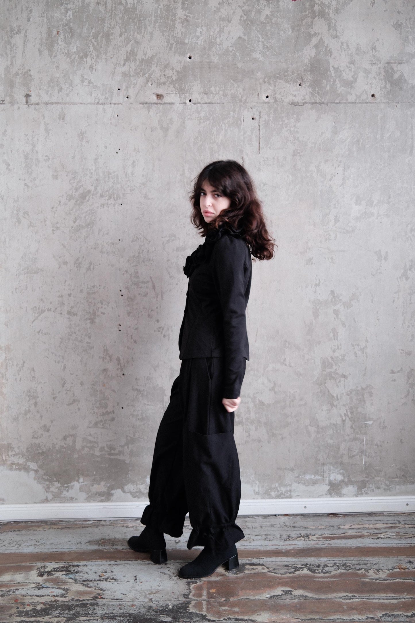 A woman in a black outfit stands in a room with a concrete wall.