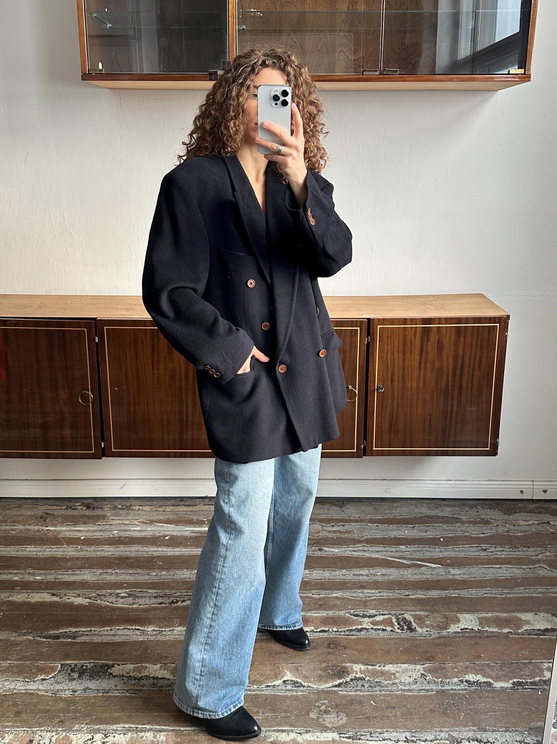 A person is taking a selfie with a black blazer and blue jeans, standing in front of a wooden cabinet.